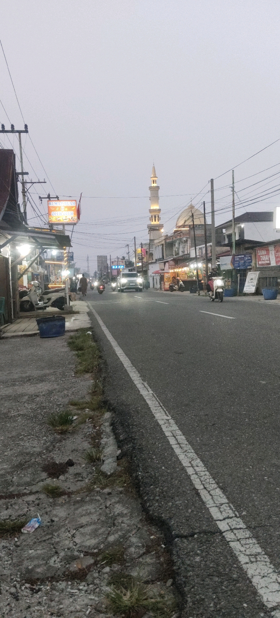 The image depicts a street scene in Sungai Pakning, Indonesia, likely captured during twilight or overcast conditions due to the diffused natural light. A prominent minaret of a mosque, illuminated by artificial lights, rises in the background, alongside the building's dome. The street is paved and features a white dashed line marking the lane.
On the left side of the road, a sidewalk area is visible, with several parked motorcycles and a small roadside stall or shop with its lights on. A blue plastic trash bin is placed near the edge of the sidewalk. Further down the street, commercial buildings with illuminated signage line the road. One sign displays "RUMAH" and "Oleh-oleh", suggesting a souvenir shop or local specialty store.
Several vehicles are present on the road, including a white SUV driving away from the camera, and multiple motorcycles, some with riders. A pedestrian is seen walking on the side of the road on the left. The environment appears to be an urban or semi-urban setting with utility poles and overhead wires crisscrossing the sky. The general condition of the sidewalk suggests some wear and tear, with patches of grass growing through cracks. A discarded juice box can be seen on the cracked pavement of the sidewalk in the foreground.