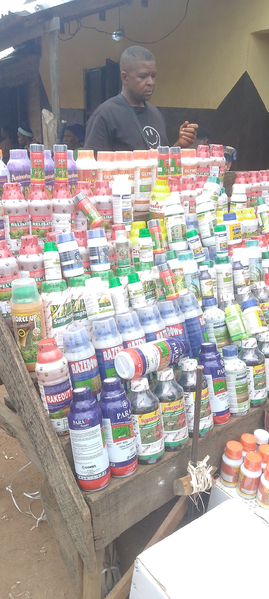 The image depicts an outdoor market scene in Tor-Donga, Nigeria, likely a street stall or roadside vendor. A Black male, middle-aged, wearing a black t-shirt with a smiley face logo, stands behind a display of various agricultural products, primarily bottles and containers of herbicides, pesticides, and fertilizers. The products are stacked in an organized, albeit dense, manner on wooden planks and shelves. The background shows a simple structure, possibly a stall or part of a building, with a yellow-brown painted wall and a corrugated metal roof. The ground appears to be dirt or gravel. Other individuals are partially visible in the background, suggesting a populated commercial area. The lighting suggests it is daytime. The overall impression is one of commerce and local trade.