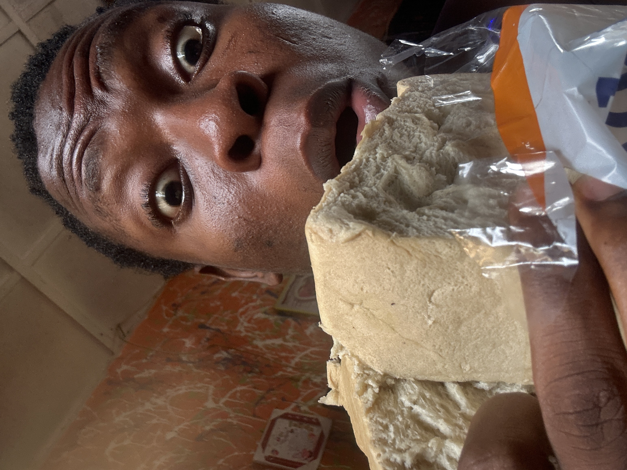 A low-angle close-up shot shows a person with dark skin and dark, curly hair, with a large block of bread held close to their open mouth. The person's eyes are wide, and their head is tilted. The bread is wrapped in clear plastic, and the packaging has orange and white stripes. The background consists of a surface with an orange and white patterned covering, and a glimpse of a tan wall. The location is Unknown, Unknown.