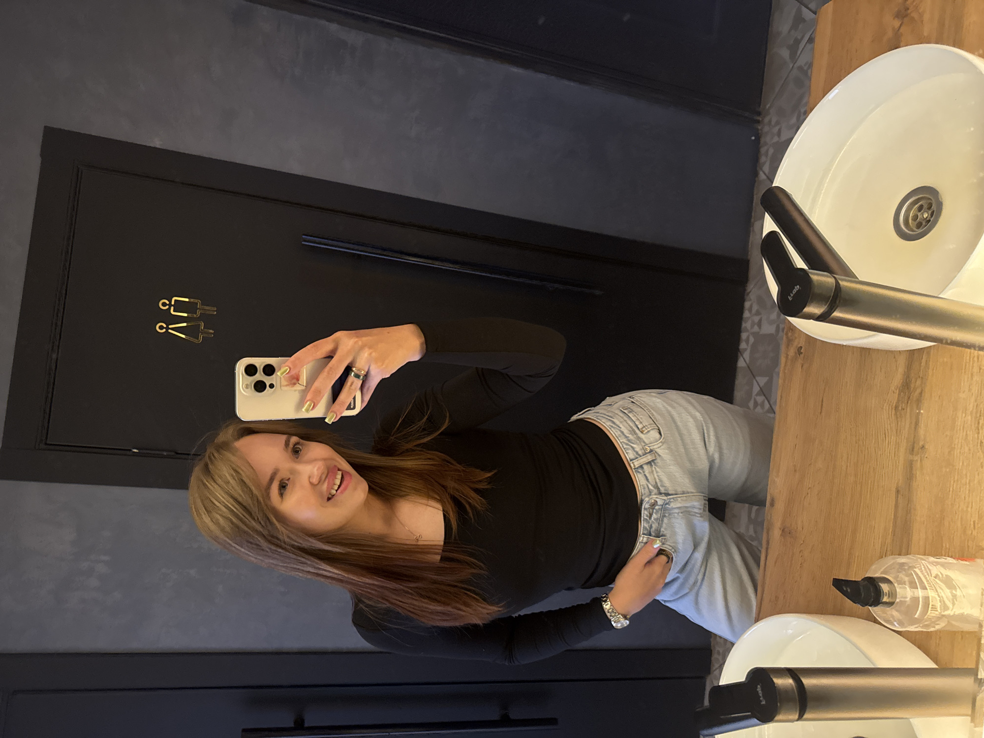 A woman with long, light brown hair, wearing a black long-sleeved shirt and light blue jeans, takes a mirror selfie in a dimly lit restroom. She is smiling and holding a smartphone in her right hand, displaying bright green nail polish. Her left hand is in her pocket, and she wears a watch on her left wrist and a ring on her left index finger. Behind her, a black door is visible with a gold symbol depicting a man and a woman. The restroom features dark gray walls, a wooden countertop, a white sink with a chrome faucet, and a bottle of hand soap. The environment suggests a modern, stylish public restroom. The specific location in Chaikovsky, Russia, is not discernible from the image itself.