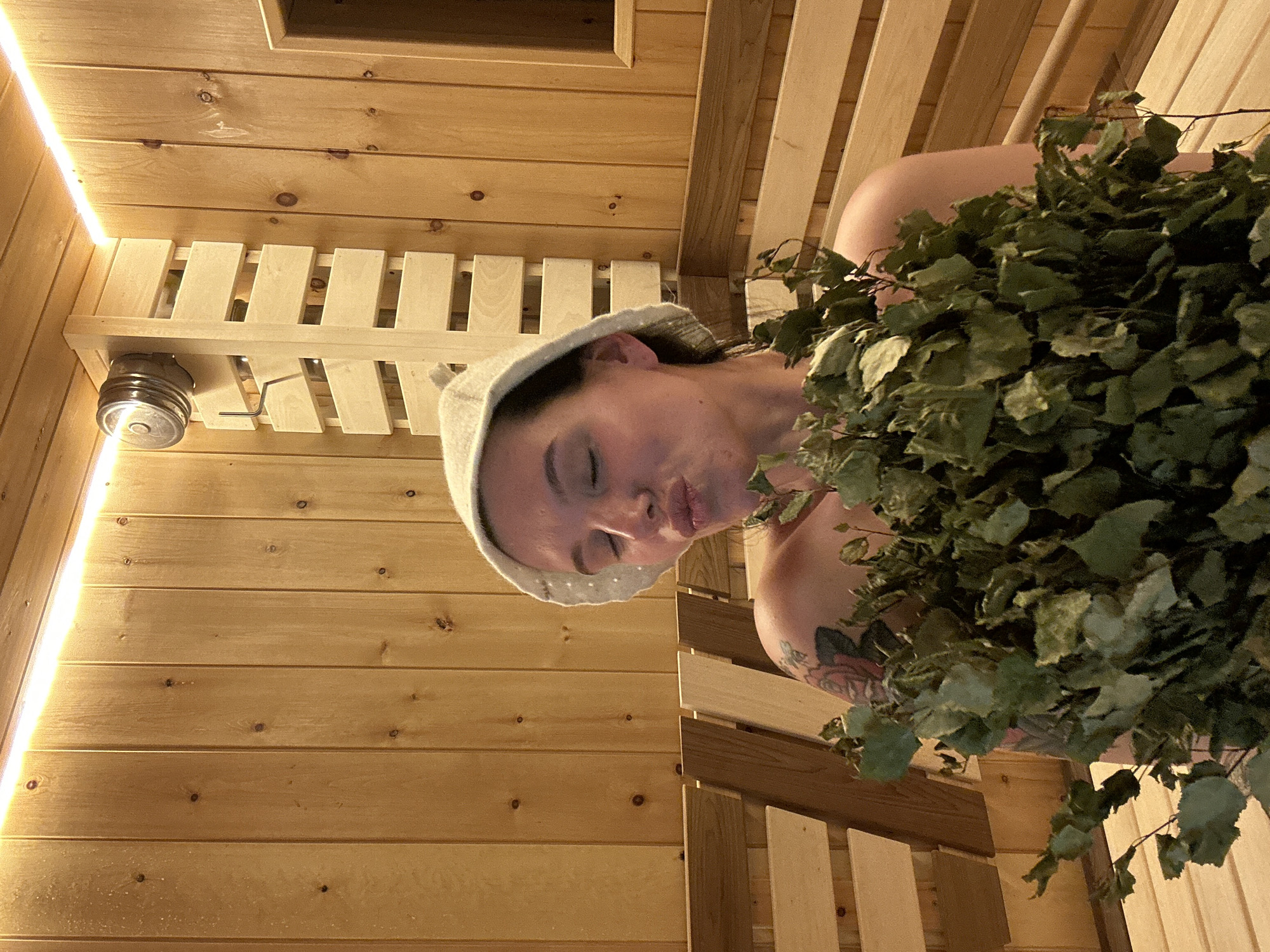 The image appears to be taken inside a wooden sauna, likely located in Прикамский, Russia, judging by the typical sauna culture. A woman is sitting on a bench, wearing a sauna hat. She holds a bundle of leafy branches, often used in saunas for therapeutic purposes. The sauna interior consists of light-colored wooden planks. There is a light fixture on the upper left.
