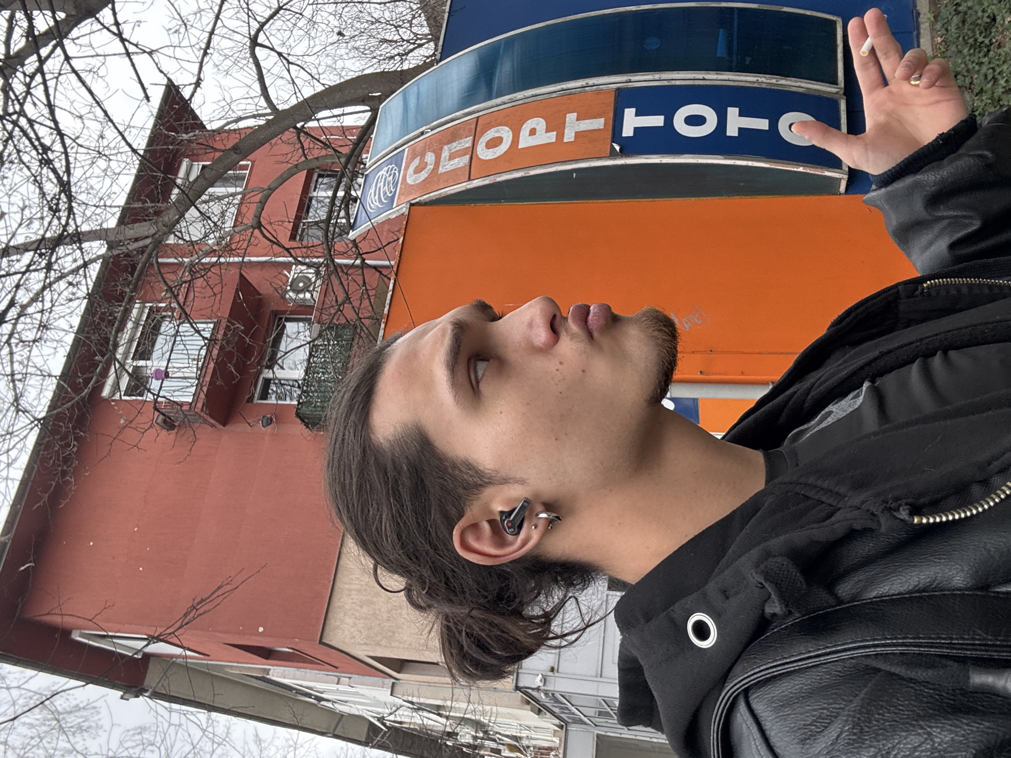 A man is positioned in front of the building in Sofia, Bulgaria, identified by a sign displaying "SPORT TOTO". He is facing upwards, holding a lit cigarette between his fingers. The man has dark hair, partially tied back in a bun, with a goatee, and wearing an earpiece. He is dressed in a black leather jacket. The background includes a red building with visible windows and bare tree branches.