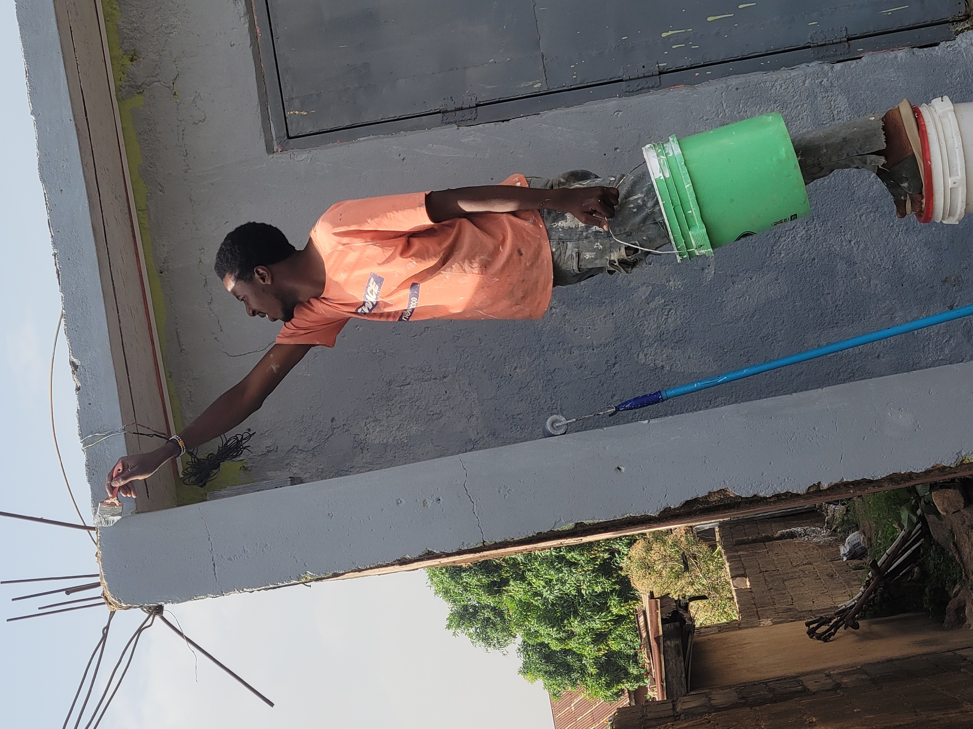 An outdoor image, captured in bright daylight under a clear sky, depicts a man painting a building in what appears to be a construction or renovation setting in Jos, Nigeria. The man, with dark skin and short dark hair, is seen in profile, wearing an orange t-shirt with the text "VICE" and "supreme" partially visible, and camouflage pants. He is focused on painting the edge of a concrete structure with a small paintbrush, his left arm extended.
A green plastic bucket is suspended from his left hip, and he appears to be standing precariously on a white plastic bucket, which itself rests on an unseen support, enabling him to reach the elevated section. The building wall is a light gray, with patches of yellow primer or old paint visible. Exposed rebar protrudes from the raw concrete edge of the structure, indicating ongoing work. A paint roller with a blue handle is visible further along the wall.
Below the painted structure, lush green trees and foliage are seen, along with a rustic ground area containing stacked wood or debris. A dark, possibly shuttered, window or opening is on the wall above the man. The scene suggests manual labor under sunny conditions.