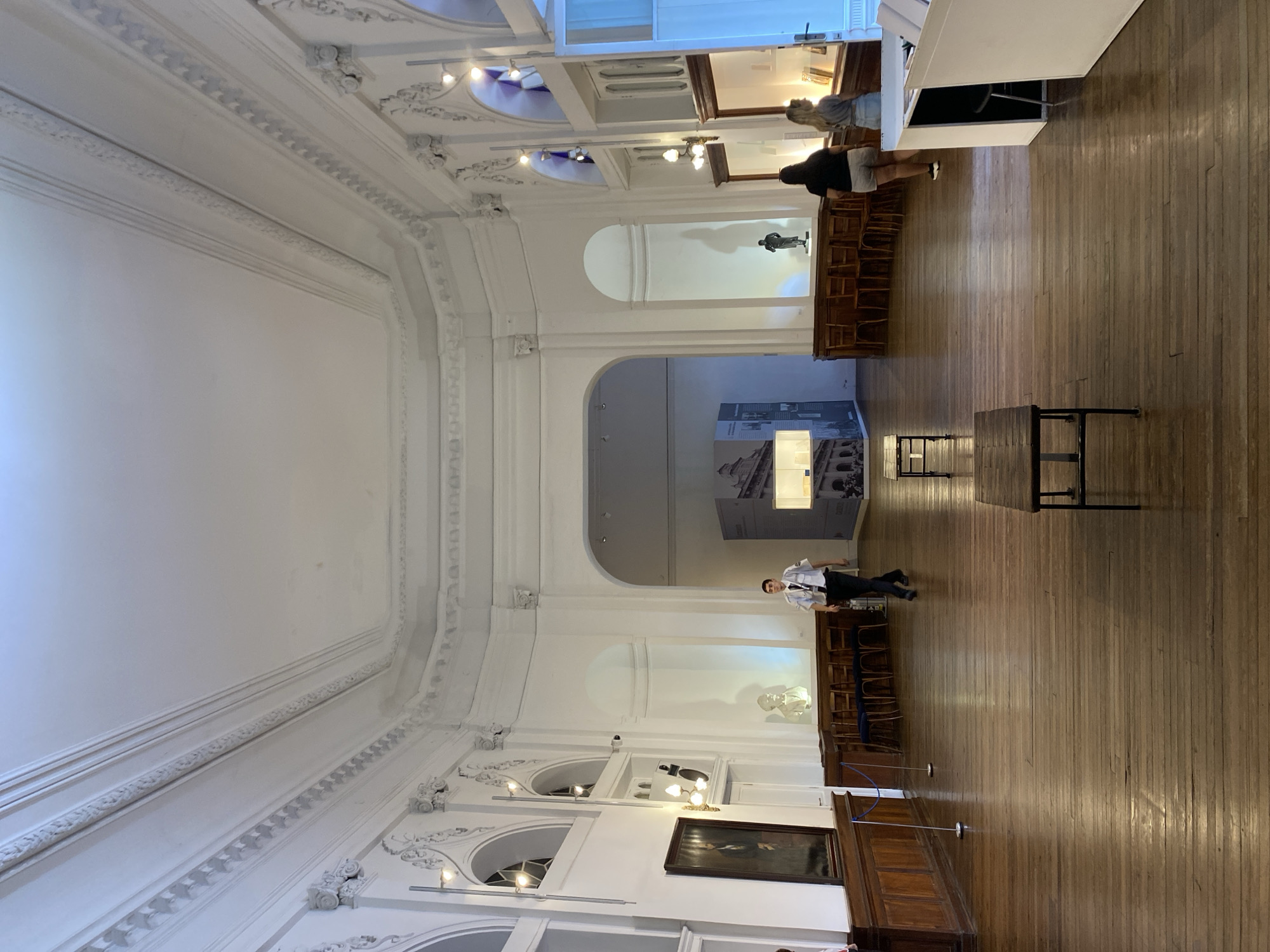 This is an interior view of what appears to be a museum hall or gallery, likely located in Buenos Aires, Argentina. The space has high ceilings with elaborate white plasterwork and architectural details. Natural light streams through arched windows along one side, illuminating a polished wooden floor.
Several individuals are present:
* One person dressed in what looks like a pilot's uniform is near the middle of the hall, possibly walking.
* Two additional people are located at the top of the photo.
Various objects and elements are also visible:
* Artwork, including a framed portrait, is displayed along the walls.
* Wooden benches line the sides of the hall.
* A screen or display case with an image or exhibit is visible at the far end of the hall.
* A couple of small tables are placed in the room.