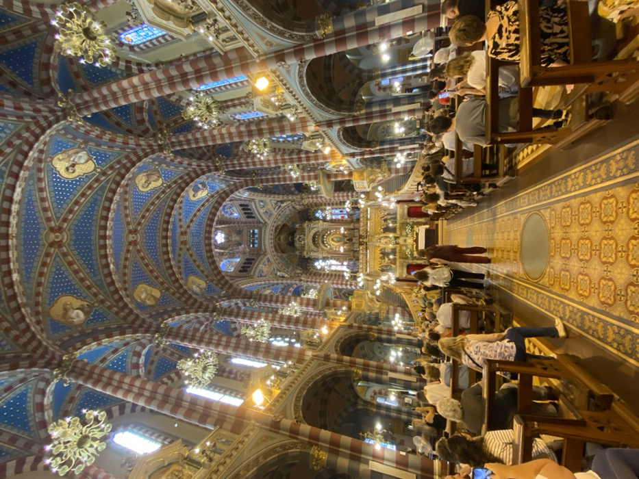 The image displays the interior of the Basílica Nuestra Señora del Pilar, located in Recoleta, Buenos Aires, Argentina. The perspective is from the rear of the nave, looking directly towards the ornate main altar.
The basilica features a high, vaulted ceiling extensively painted in dark blue with gold stars and circular medallions depicting religious figures. Supporting this ceiling are numerous columns and arches, all uniformly striped horizontally in alternating red and light grey/white bands. Multiple ornate golden chandeliers hang from the ceiling.
At the far end, the main altar is an elaborate, multi-tiered gilded structure, elevated and flanked by symmetrical grand staircases. A prominent statue is visible in a central niche on the altar, surrounded by other statuary and decorative elements. White floral arrangements are placed near the altar. The central aisle is covered by a patterned floor runner in shades of brown and gold.
Approximately 50 to 70 individuals, primarily adults of varying ages, are seated in dark wooden pews on both sides of the central aisle, facing the altar. Several individuals are observed holding up smartphones, seemingly recording or photographing the scene. The overall lighting is a blend of artificial light from the chandeliers and natural light filtering through stained-glass windows in the upper sections of the walls.