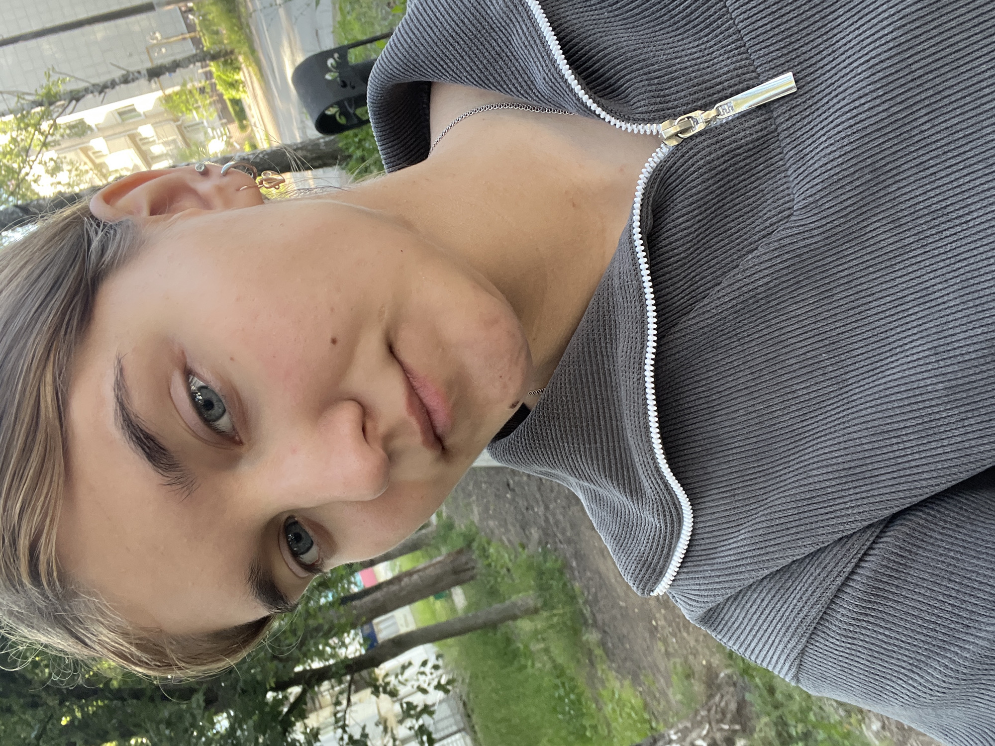 The image is a close-up, rotated selfie of a young person with fair skin and blue eyes, taken outdoors. They are wearing a dark grey, ribbed zip-up jacket and a delicate silver necklace. Visible piercings include a stud in the upper ear and a gold hoop earring. The background shows greenery and trees, suggesting a park or outdoor public space. Buildings and a street are faintly visible in the distance, indicating an urban or suburban environment. The lighting suggests it is daytime, possibly late afternoon due to the soft light. The person's expression is neutral. No visible text is present in the image. The location context is Chaikovsky, Russia.