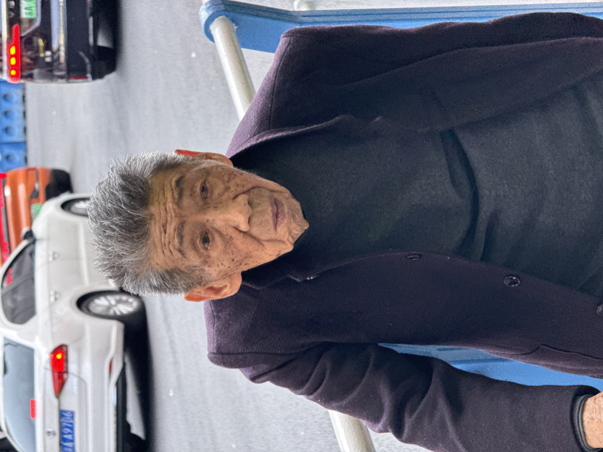 An elderly man with gray hair and wrinkled skin is seated in what appears to be a blue-framed chair or bench. He is wearing a dark-colored jacket or sweater over a dark top. In the blurred background to the left are several vehicles including what looks like a white car with the license plate "浙A A9706", and what looks like a black and an orange car next to it. Due to the license plate being shown, this may be taken in the Yuzhong District of China. The focus is on the man, capturing his aging features and the texture of his clothing.