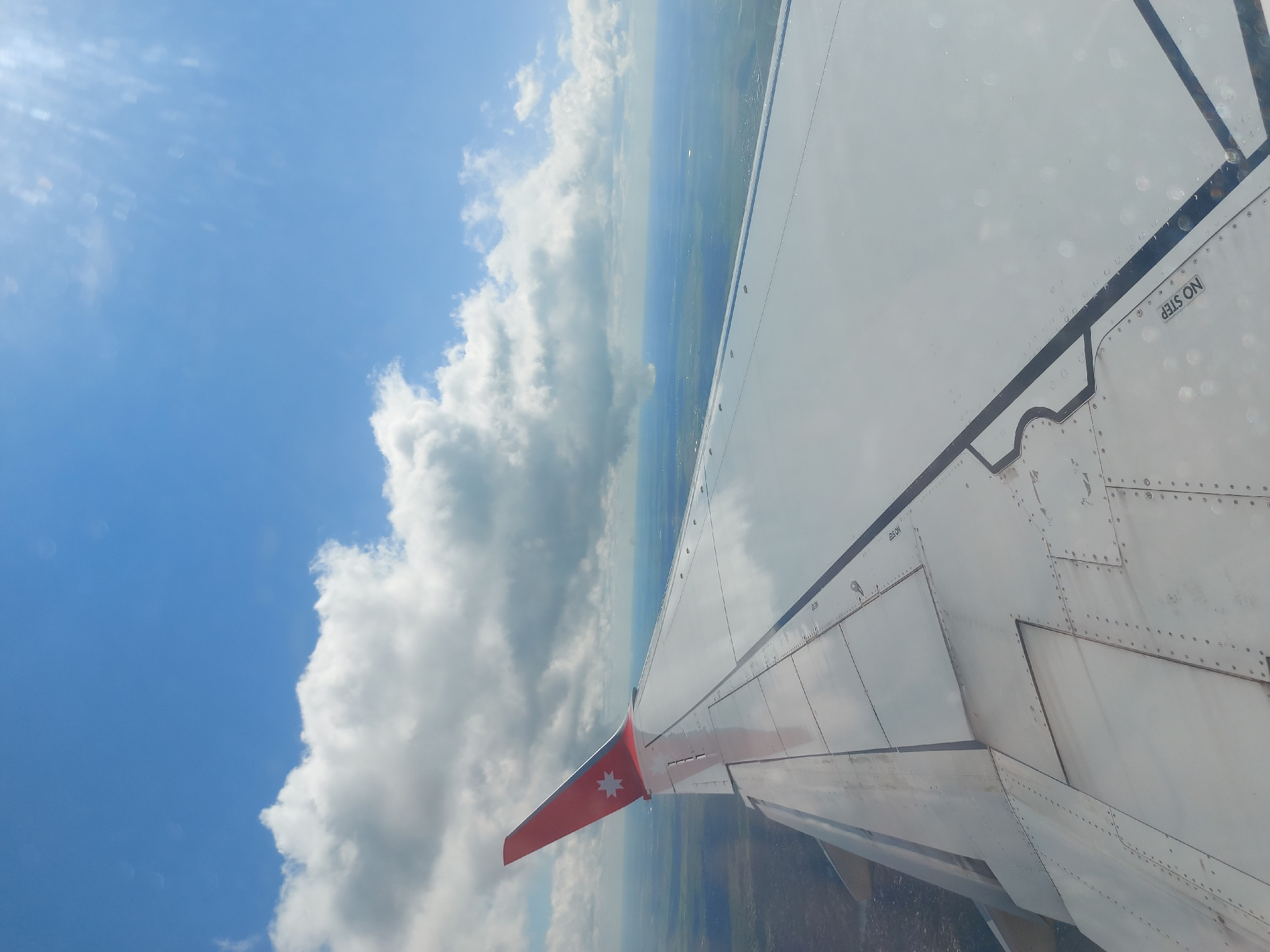 The image shows the wing of an airplane in flight. The wing is mostly white, with a red-painted wingtip. The sky is blue with scattered white clouds to the left. Below the wing and clouds, the ground appears to be a mix of green fields and possibly forest. The words "NO STEP" are visible on the wing. It is not possible to verify the location of Новое Мартьяново, Russia from the given image.