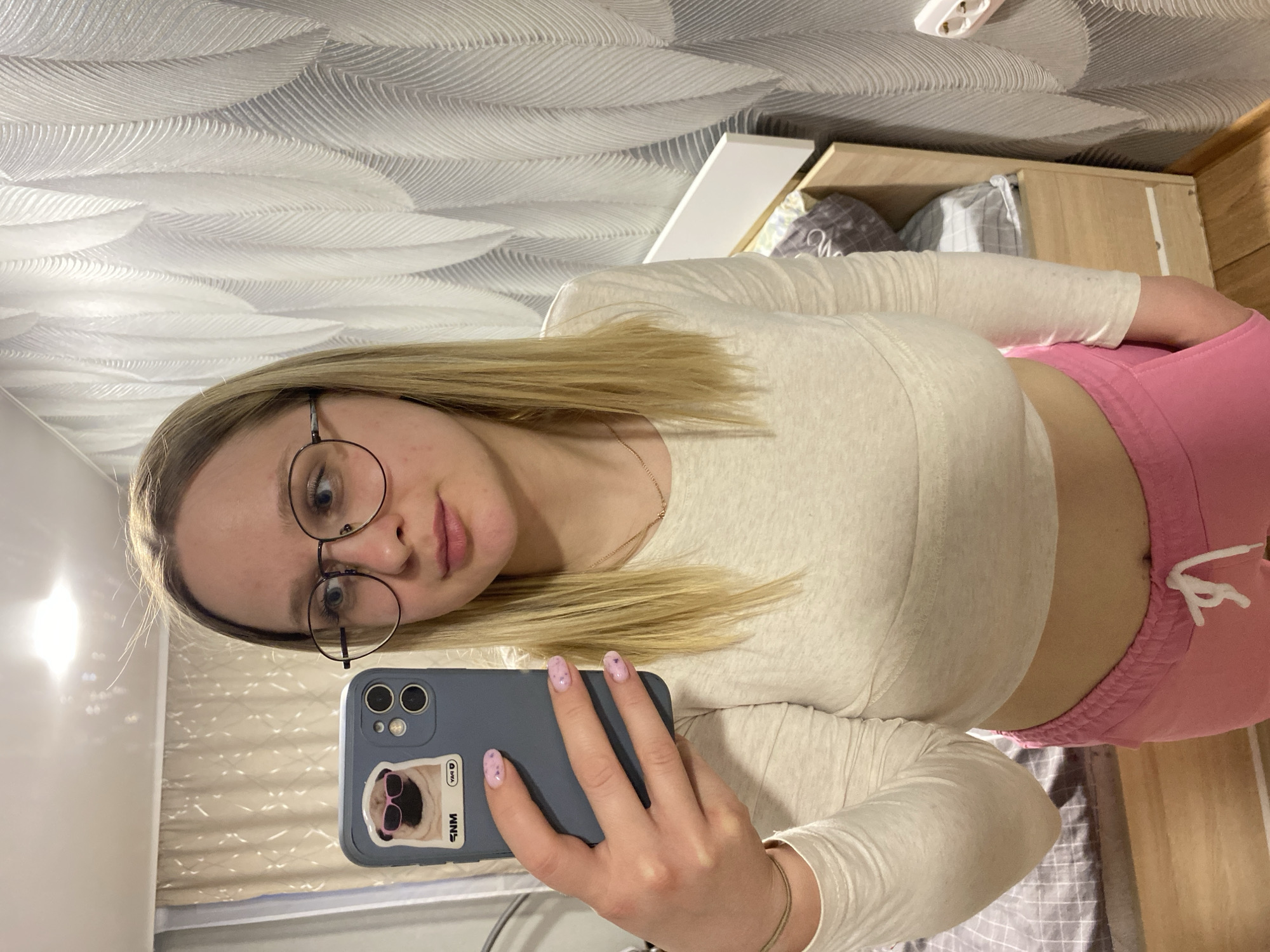 A woman with blonde hair and glasses is taking a selfie indoors. She is wearing a long-sleeved off-white shirt and pink pants. The phone case she's using is blue and has a sticker of a dog on it. The background shows a bed and a patterned wall. The location is presumed to be indoors, possibly a bedroom. There is no information provided to suggest this is in Izhevsk, Russia.