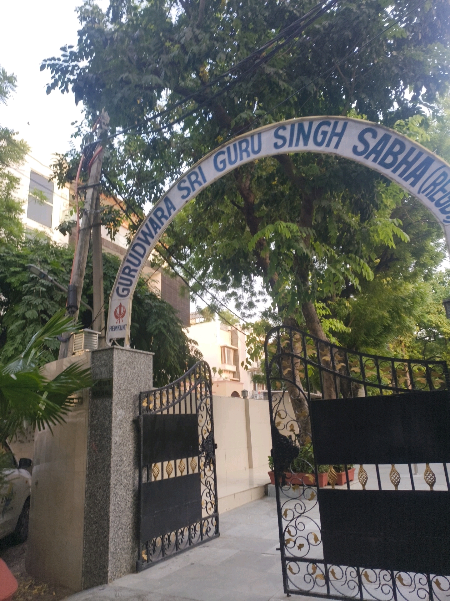 The image captures the entrance to Gurudwara Sri Guru Singh Sabha, located in Delhi, India. The primary subject is an arched signboard above the entrance, with the text "GURUDWARA SRI GURU SINGH SABHA (REGD.)" inscribed in blue letters. Below the arch, a pillar displays the Sikh Khanda symbol and the word "HEMKUNT" in black text. The entrance features a black wrought-iron gate with ornate golden detailing, which is partially open, revealing a pathway and potted plants. To the left of the gate, a portion of a building with a tiled exterior is visible, along with some foliage. A white car is parked to the far left, partially obscured by a palm frond. The environment appears to be outdoors, with trees providing shade and a slightly overcast sky. The overall scene depicts a religious institution entrance in an urban setting.