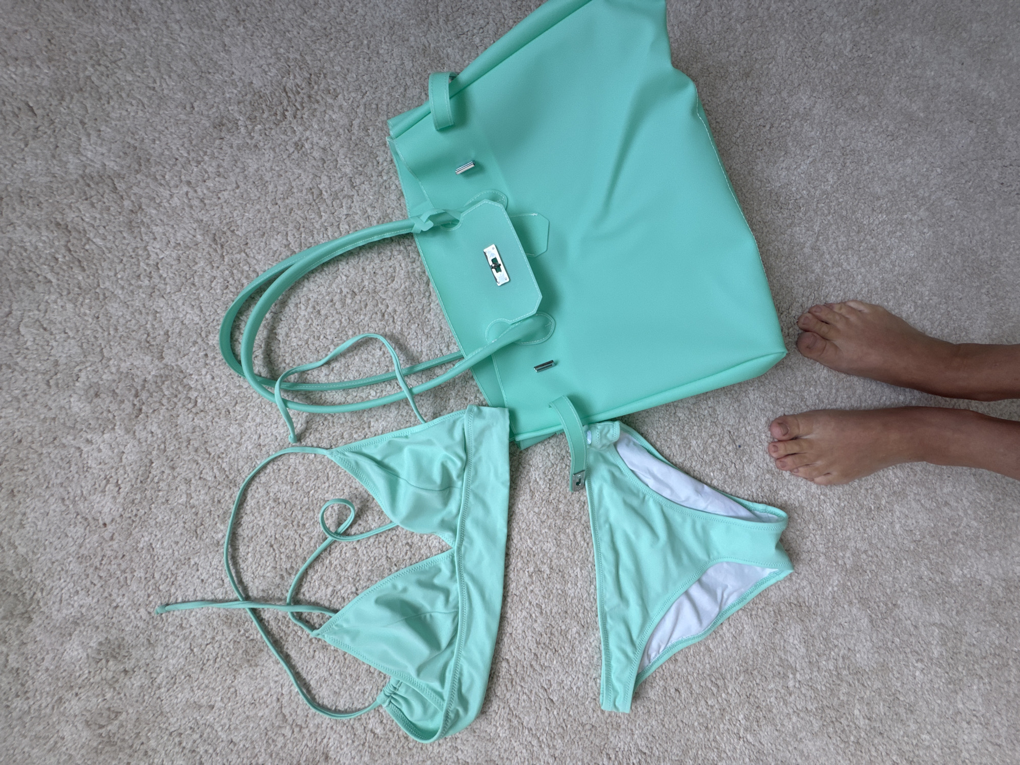 A flat lay image shows a turquoise bikini and a matching turquoise tote bag on a beige carpet. The bikini top is a triangle style with thin straps, and the bottoms have a similar style with a buckle detail on the side. The tote bag is large with two handles and a clasp closure. Two feet, belonging to a person, are visible in the foreground. The image appears to be taken indoors, possibly in a home. There is no indication of the location being in Анкудиновка, Russia.