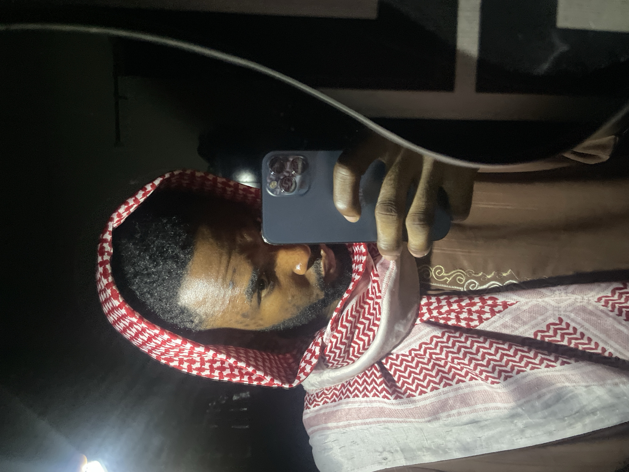 A person is holding a smartphone up to a reflective surface, likely taking a selfie. The person is wearing a traditional red and white checkered headscarf (keffiyeh) and a brown robe or shirt. The reflection shows the person's face. The environment is dark, with some light sources obscured by the reflective surface. Location information is not discernible from the image.