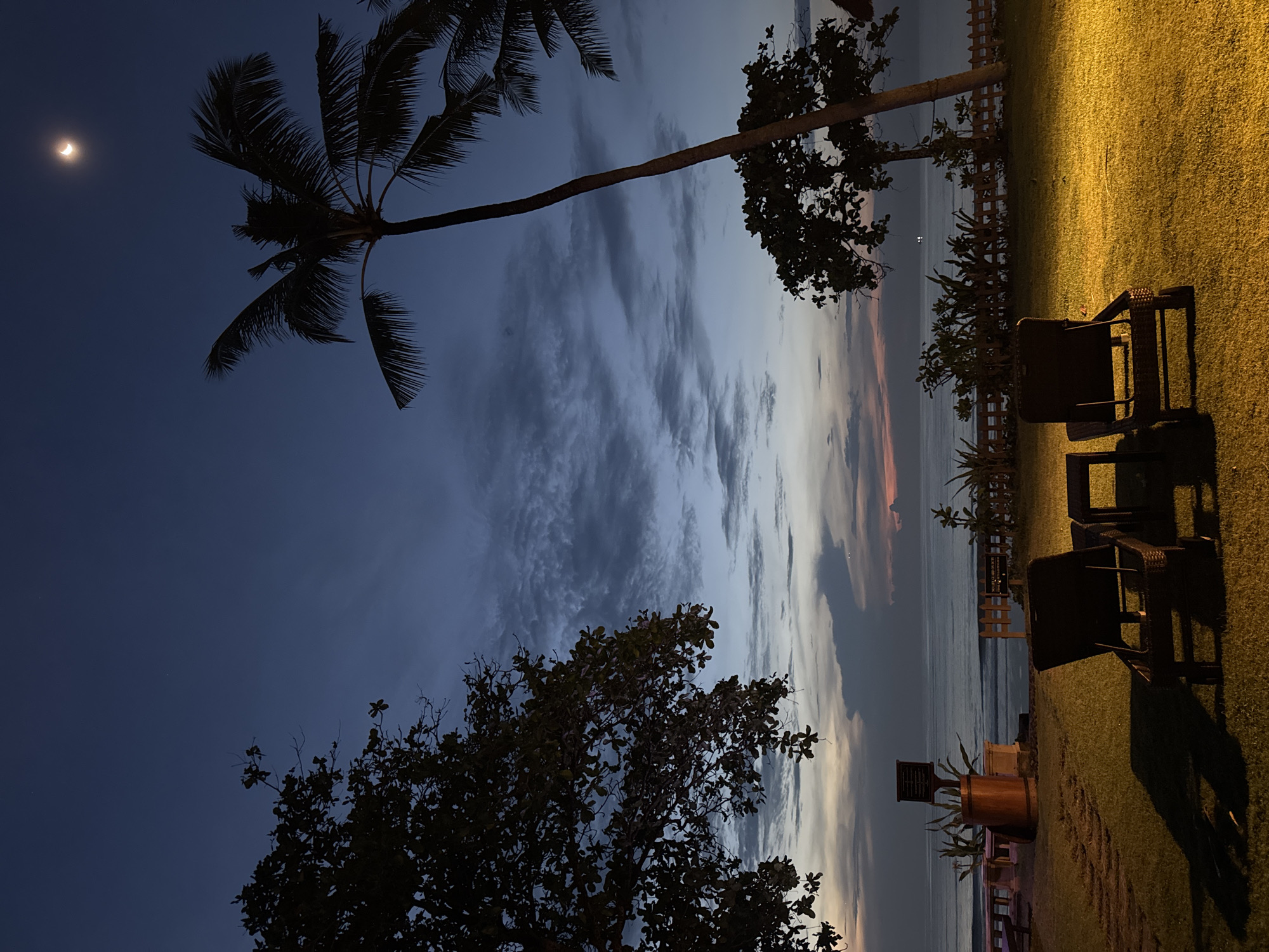 The image depicts an outdoor scene in Kuta Selatan, Indonesia, at dusk or night. The sky is a gradient of dark blue to shades of pink and light grey, with scattered clouds. A crescent moon is visible in the upper-left portion of the frame. Silhouetted palm trees and other trees frame the sky. In the foreground, there is a grassy area, illuminated by artificial light that casts long shadows. Two dark-colored wicker chairs and a small table are positioned on the grass, overlooking what appears to be a body of water, likely the ocean. A wooden fence and some vegetation are visible near the edge of the grass. Further to the left, on the grass, there is a barrel-like structure. The setting suggests a resort or private property near the beach.