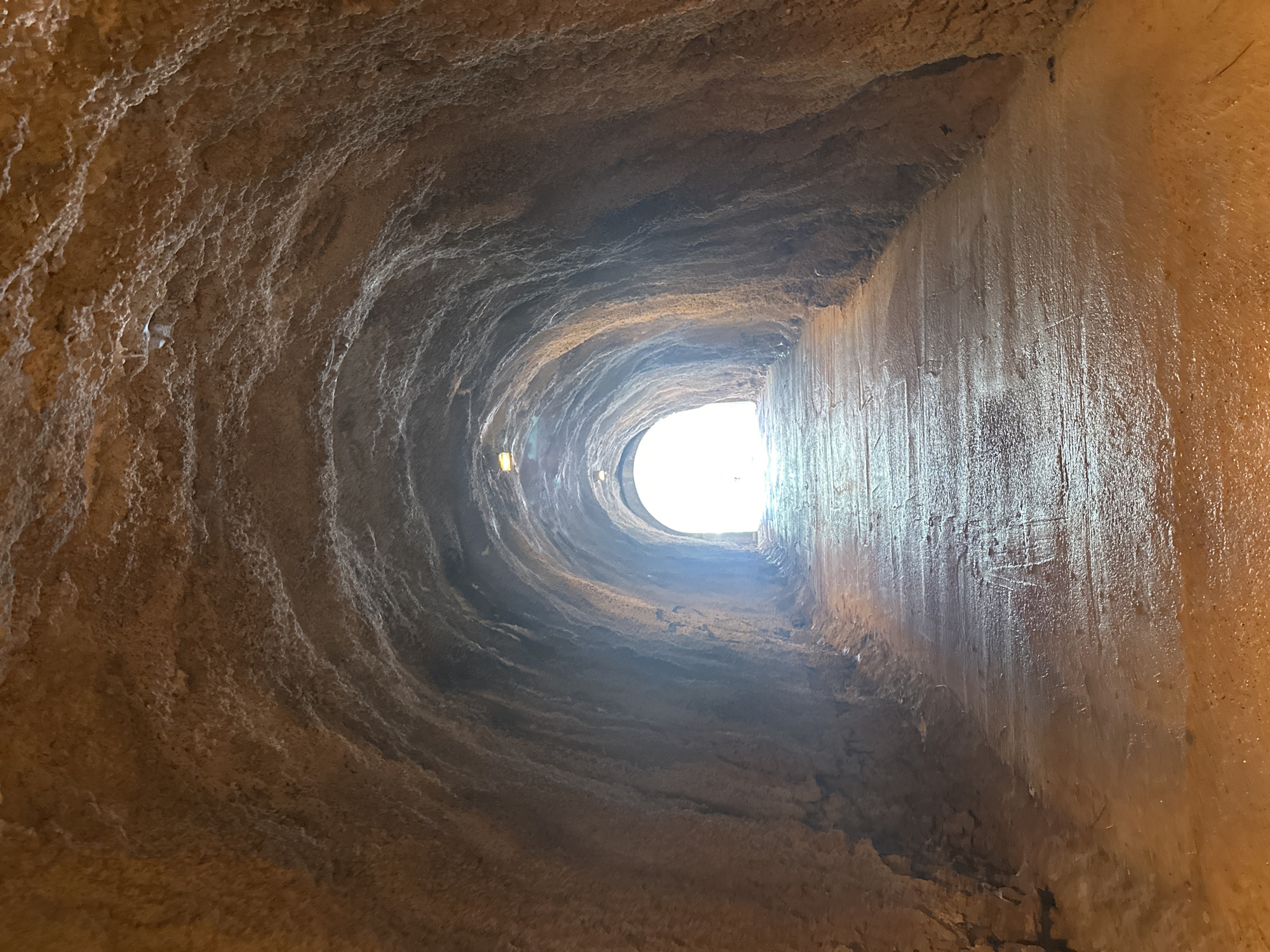 The image shows the interior of the abandoned water channel of Kamenovo, Montenegro. The water channel is a rough-hewn, curving tunnel of pale brown rock and concrete. The left side of the tunnel is curved and porous. The right side of the tunnel is a flat concrete surface with water damage. Light enters the tunnel at the tunnel's end, which is a round hole. The only light source is from the tunnel end and the reflection from the channel walls.