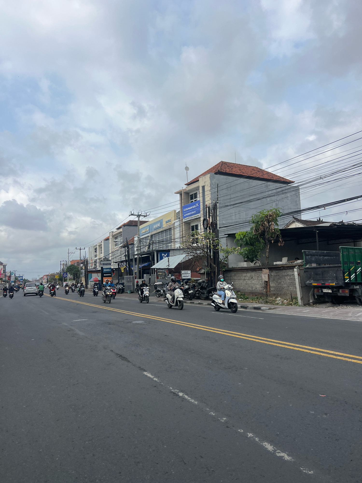 The image depicts a street scene in Denpasar, Indonesia, under a cloudy sky. The road, marked with yellow double lines, is busy with traffic, primarily motorcycles. Several individuals are riding motorcycles, some with passengers. On the right side of the road, there are buildings with shops and businesses, including one with a "Goodyear" sign. A large truck is parked on the right side. The architecture suggests a mix of commercial and residential structures. The environment is urban, with power lines crisscrossing overhead. The general atmosphere appears to be that of a typical day in a bustling Indonesian city.