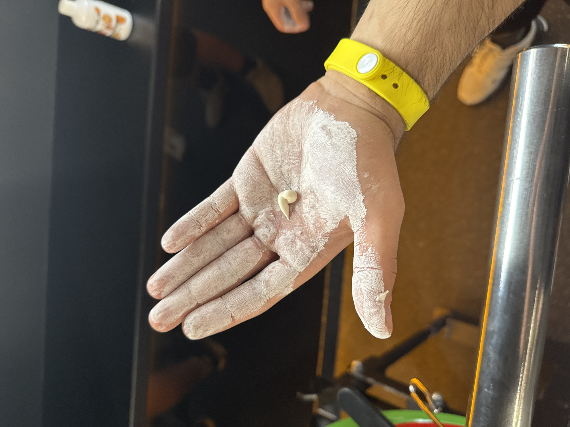 A close-up vertical shot displays a person's right hand, palm facing upwards and heavily coated with a white, powdery substance, consistent with gym chalk. A small, pale, off-white object, approximately 1-2 centimeters in length and curled into a loose spiral or "J" shape, rests in the center of the chalk-dusted palm. A bright yellow wristband with a circular white button is worn on the person's right wrist, partially obscured by arm hair. The background is largely out of focus, showing a dark, indistinct environment that suggests an indoor facility. A metallic, cylindrical bar runs across the lower foreground. In the upper right background, a blurry white bottle with an orange label is visible on a dark surface. Other indistinguishable shapes, possibly equipment and footwear, are visible in the blurred mid-ground. The overall lighting is subdued, with artificial illumination. Location: Unknown, Unknown.