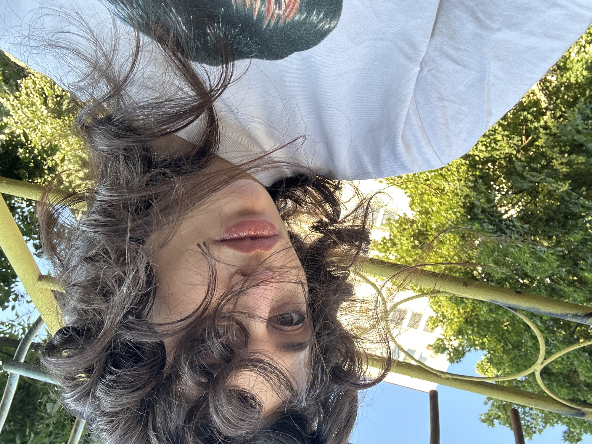 The media file is a close-up, upside-down selfie of a young person with dark, curly hair. They are wearing a white t-shirt with a dark graphic on it. Their face is turned towards the camera, and their eyes are open, looking directly at the viewer. They have light-colored skin and full lips that appear to be glossed. The background is blurred, but hints of green foliage and a bright blue sky suggest an outdoor setting, likely during the daytime under fair weather. There are also visible elements of a yellow railing or fence in the foreground. The overall impression is casual and candid, possibly taken during a moment of leisure outdoors. The location context provided places the scene in Sofia, Bulgaria. No visible text is present in the image.