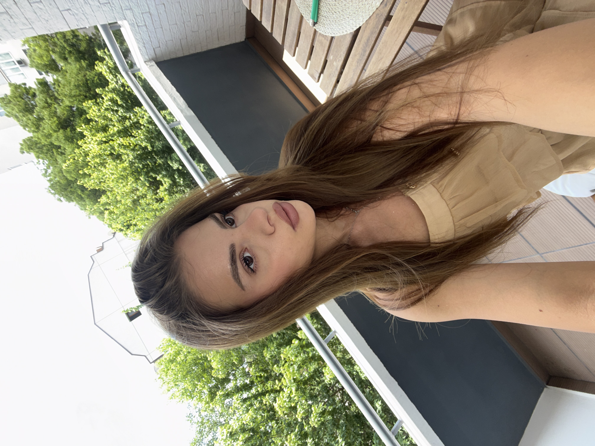 This is a daytime close-up selfie of a young woman, taken on what appears to be a balcony or outdoor patio in Plovdiv, Bulgaria.
The main subject is a young woman, positioned at an angle, looking directly at the camera. She has long, straight brown hair that frames her face and flows over her shoulders. Her complexion is fair, and she has defined eyebrows, brown eyes, and full, slightly parted lips with a neutral or subtly pouting expression. She is wearing a light brown or beige garment, possibly a dress or top, with pleats visible on the shoulder area, and a thin silver necklace.
The setting is an outdoor space, likely a balcony, indicated by a metal railing and dark grey wall to the woman's right. Beyond the railing, vibrant green trees are visible, suggesting a natural or park-like area in the background, along with part of a white building. To her upper right, a wooden slatted surface, possibly a bench or table, holds a light-colored straw hat, on which a small green object (possibly a lighter) rests. The lighting is bright and sunny, indicating clear weather during the day. There is no visible text in the image.