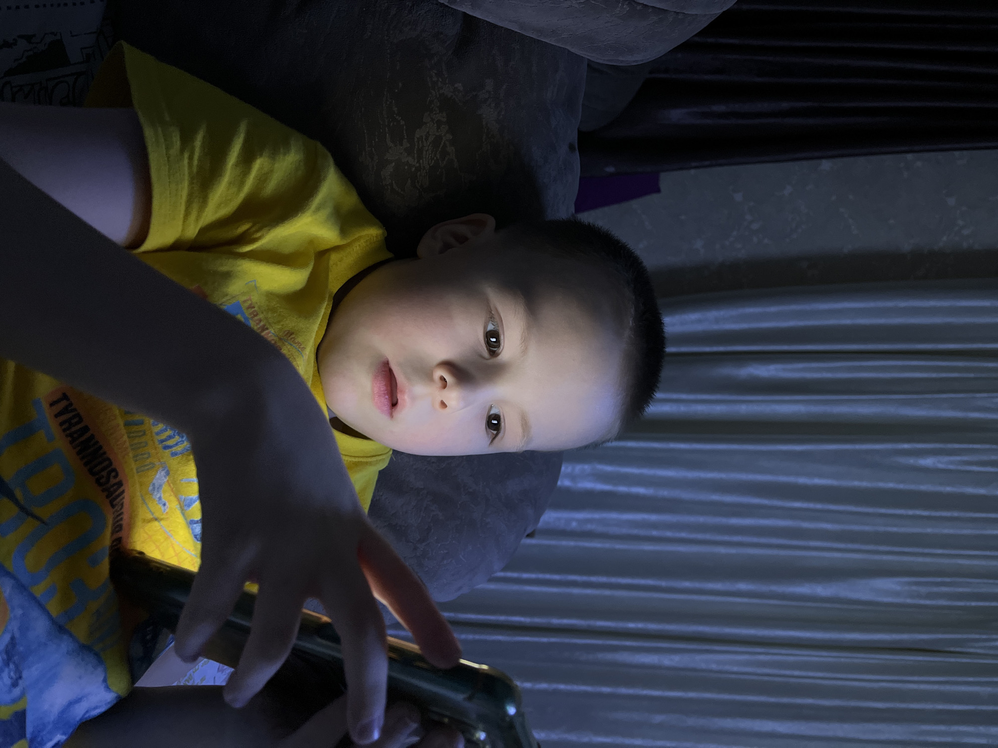 A young child in a yellow "TYRANNOSAUR" t-shirt is lying down, possibly on a sofa, holding and looking at a mobile phone. The lighting suggests the room is dimly lit, with the phone screen illuminating the child's face and hand. The background features a dark curtain or drape. The image does not provide enough information to confirm its location in the city of Usman, Russia.
