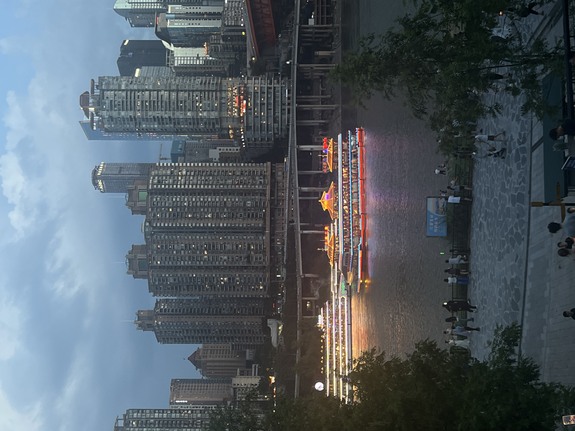 The image depicts a cityscape at dusk, likely in Jiangbei Cheng Street, China. In the foreground, a cobblestone plaza is populated by several individuals. Some are standing and conversing, while others appear to be walking. A large, dark green tree dominates the left side of the frame, obscuring some of the view.
The middle ground features a body of water, possibly a river, on which two illuminated tour boats are navigating. The boats are adorned with colorful lights, reflecting on the water's surface. A third, larger boat with extensive lighting is visible further in the distance, on the right side of the river.
The background is characterized by a dense collection of high-rise buildings, indicative of a modern urban environment. These skyscrapers display a variety of architectural styles and are illuminated by internal lights. Elevated highways and bridges span across the scene, connecting different parts of the city. The sky is overcast with clouds, suggesting either twilight or impending rain. The overall ambiance is one of a bustling city transitioning into the evening.