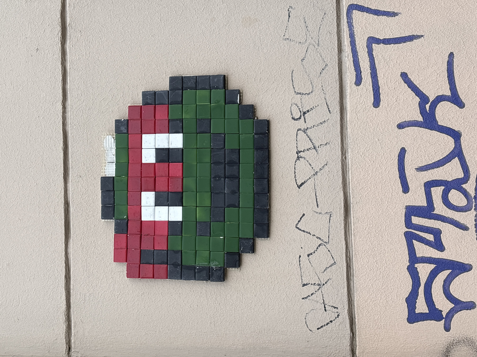 A mosaic made of small, square tiles affixed to a wall, likely an exterior wall in a public space, featuring a pixelated design of a face predominantly in red, green, black, and white. The mosaic is somewhat egg-shaped or oval. To the right of the mosaic are graffiti markings in black and blue. The wall appears to be a light, neutral color with vertical seams or divisions. There's some minor damage or missing tiles on the left side of the mosaic. The location is Mönchengladbach, Germany.