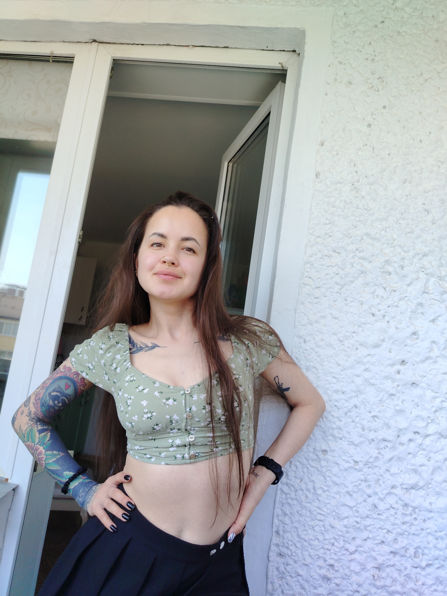 A woman with long brown hair, multiple tattoos, and piercings, stands on a balcony in Noviy, Russia. She wears a green floral crop top and a black pleated skirt. Her arms are akimbo, and she is looking at the camera. A white door frame and a heavily textured white wall are visible behind her. Sunlight illuminates the scene.