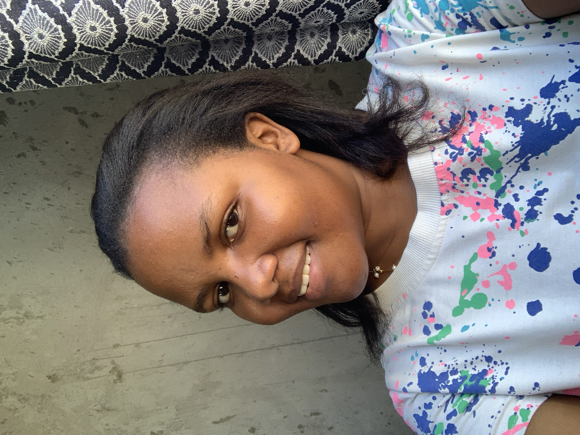 A woman with dark skin and dark hair is smiling at the camera. She is wearing a white sweatshirt with blue, pink, and green paint splatter designs. She is wearing a gold necklace with small pearls. She is lying down on a gray surface, with a patterned black and white cushion behind her. The location cannot be determined to be in Dar es-Salaam, Tanzania, based on the visual information provided.