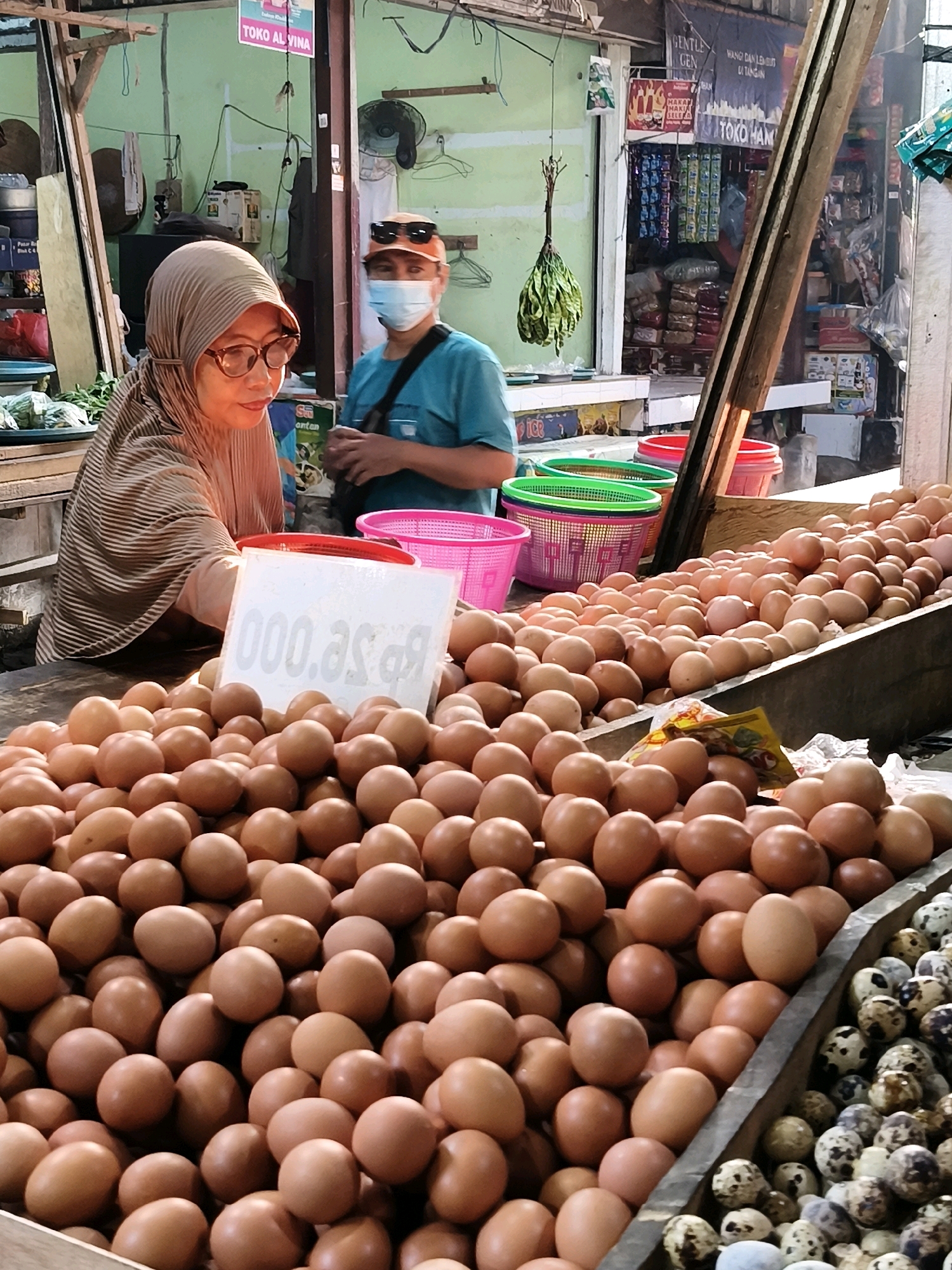 In Setiamekar, Indonesia, a market stall displays a large quantity of chicken eggs in the foreground, with smaller quail eggs to the right. A woman wearing a hijab and glasses is partially visible behind a white price sign that reads "Rp 000.asgr." To her right, a man wearing an orange hat, sunglasses, and a face mask stands near a blue t-shirt. The background reveals a market setting with shelves stocked with various goods, including packaged food items and hanging vegetables. Baskets of various colors, red and pink, are also present at the stall.