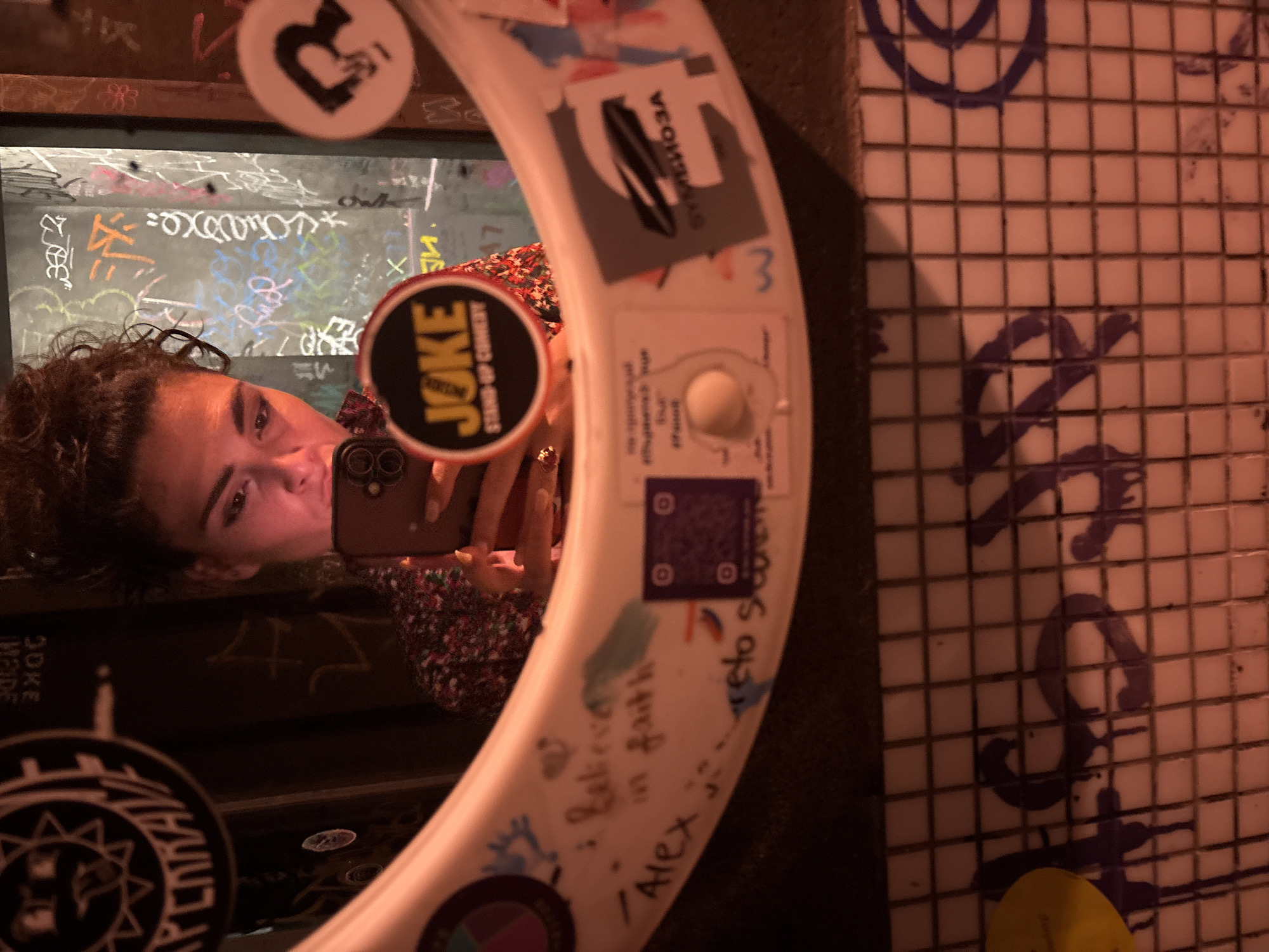 A dimly lit mirror selfie captures a woman in a restroom or bar area of what is identified as the JOKE Comedy Club in Sofia, Bulgaria. The central subject, reflected in a circular mirror, is a woman with dark, curly hair, seen from the chest up. She is looking down at a smartphone, which she holds with both hands, positioned to take the photograph. She wears a dark, patterned top and has red nail polish.
The mirror's thick, white frame is extensively decorated. On the left, a multi-colored circular sticker and handwritten text reading "I believe in faith" and "Alex is..." are visible, alongside a purple QR code sticker. Resting on the frame, held by the woman's left hand, is a prominent circular sticker for "JOKE STAND-UP COMEDY." To the right of this, another white sticker displays a black silhouette of a figure holding a microphone. At the top left of the mirror, a black circular sticker with a stylized sun-face design features Cyrillic text "СПЕНКРАЙ" (SPENKRAI). Other minor scrawls and a small white knob are also present on the frame.
The background, visible through the mirror, is a heavily graffitied wall with various tags and designs in multiple colors including white, orange, green, pink, and blue, consistent with an urban, artistic, or counter-culture venue. Below the mirror, a white tiled wall is stained with purple dripping graffiti, spelling out partial letters like "HAS" and a circular design. The entire scene is bathed in a warm, reddish-orange light, creating an intimate and slightly gritty atmosphere.