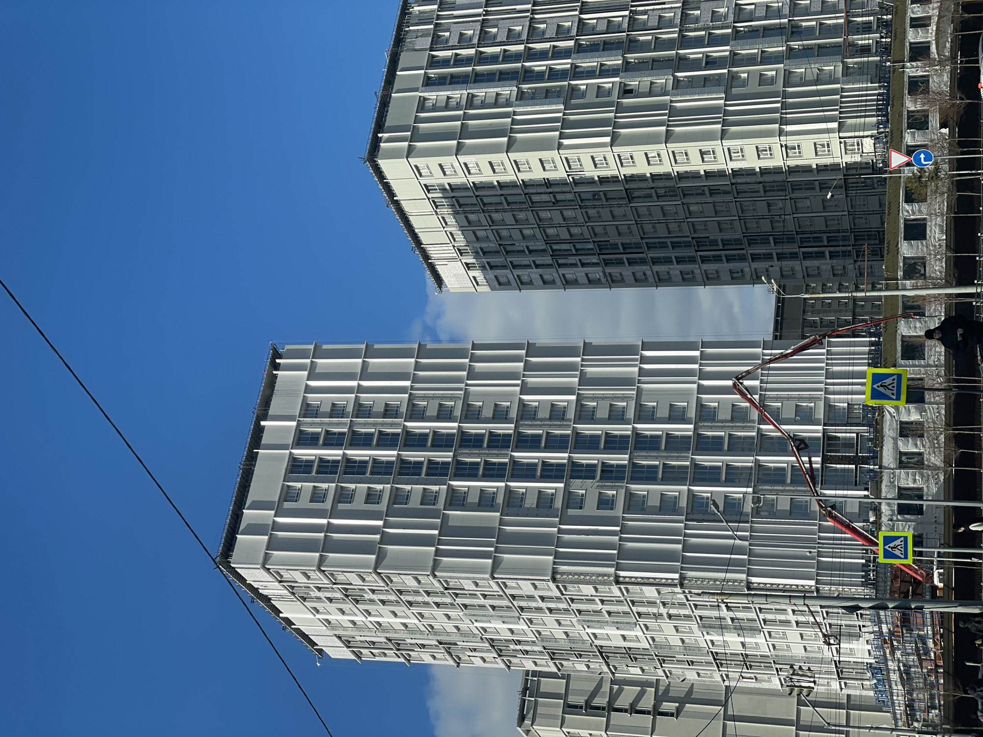 The image shows two multi-story buildings under construction in Kazan, Russia. The buildings have a repetitive window pattern and are colored gray. A concrete pump truck is visible, with its boom extended towards one of the buildings. Pedestrian crossing signs are near the buildings. The sky is blue with some clouds. A power line is in the upper left of the frame. Trees are visible.