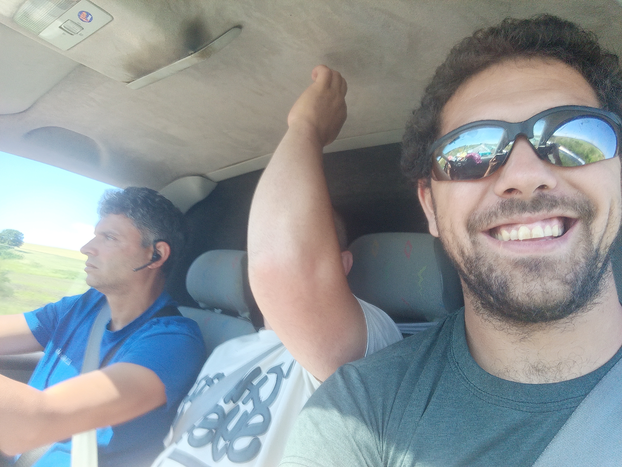 The image depicts three individuals inside a vehicle, presumably on a road trip. The primary subject is in the foreground, facing the camera with sunglasses on and a wide smile. His sunglasses reflect a road and surrounding landscape, suggesting the vehicle is in motion. To his left, another man is visible, looking ahead and appearing to be driving. He is wearing a blue t-shirt and a Bluetooth earpiece. A third person's arm is raised in the air in the middle ground, partially obscuring the view of the driver. The interior of the car shows the roof lining and seats. The scene is set outdoors during the daytime, with clear skies and a rural or suburban landscape visible through the car windows. The image does not contain any specific elements that would indicate its location in Malak Manastir, Bulgaria.