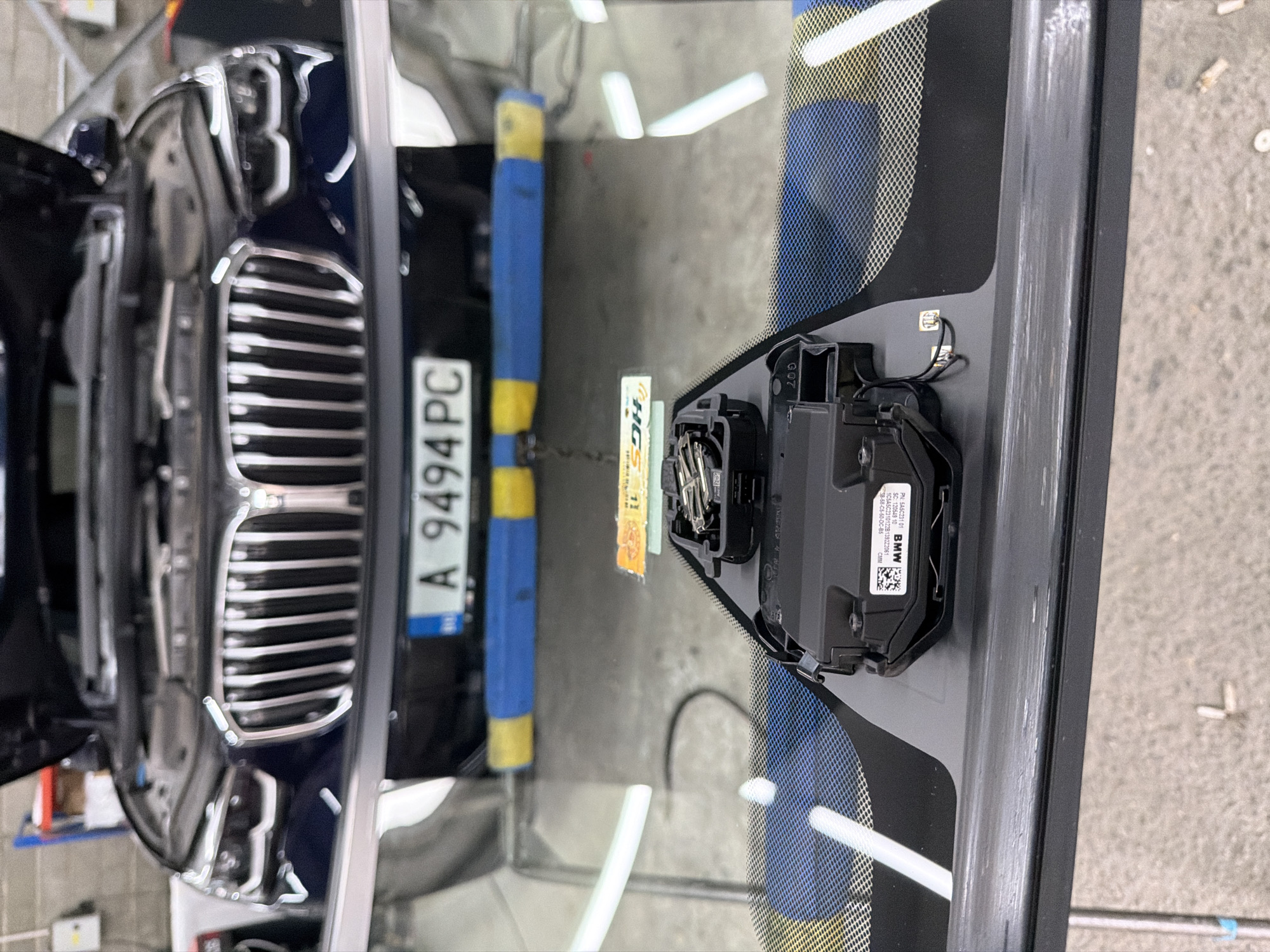 This image shows a close-up view of a car's windshield area, specifically the area where a camera or sensor module is mounted. The car appears to be dark blue or black, with its hood open, suggesting it is in a workshop or garage. A license plate with the characters "A 9494 PC" is visible on the front of the car.
The focal point of the image is a black electronic module attached to the interior of the windshield, likely for driver assistance systems. This module has a housing with visible wires and connectors. Below the module, a blue and yellow striped bumper guard is visible, possibly placed on the floor or a low platform.
The environment appears to be indoors, with a concrete floor. The background shows some shelving units and other workshop equipment, further supporting the location as a vehicle repair facility. There are no people visible in the image.
The image does not contain any information that would indicate the location is in the city of Burgas, Bulgaria.