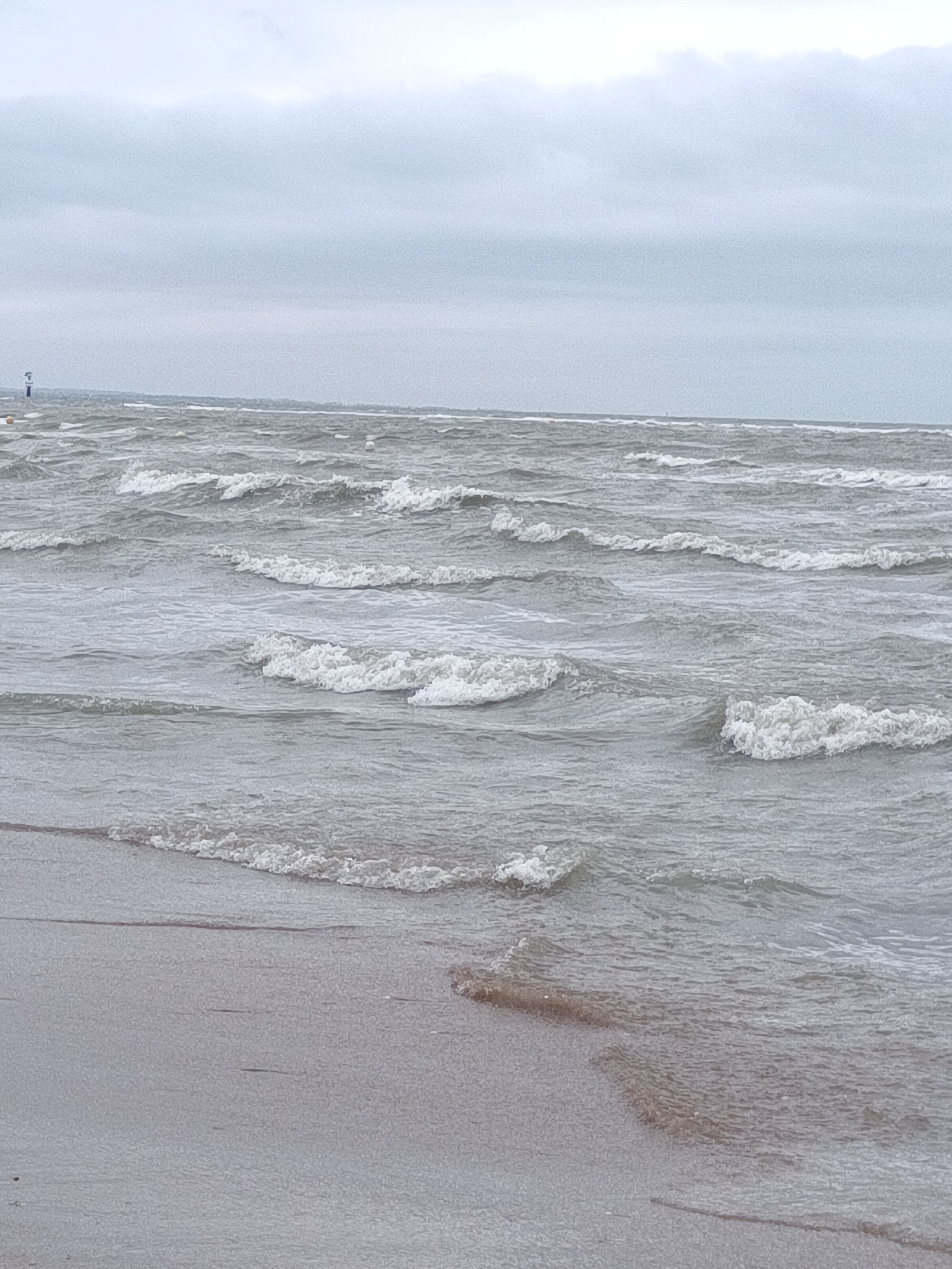 The image depicts a beach scene in Houlgate, France, under a cloudy sky. The ocean is rough, with waves rolling towards the shore. The foreground shows wet sand with ripples from the receding tide. In the distance, beyond the choppy water, a blurry horizon line is visible. There are no people or identifiable objects in the immediate foreground or mid-ground. The overall color palette is muted, dominated by grays and blues.