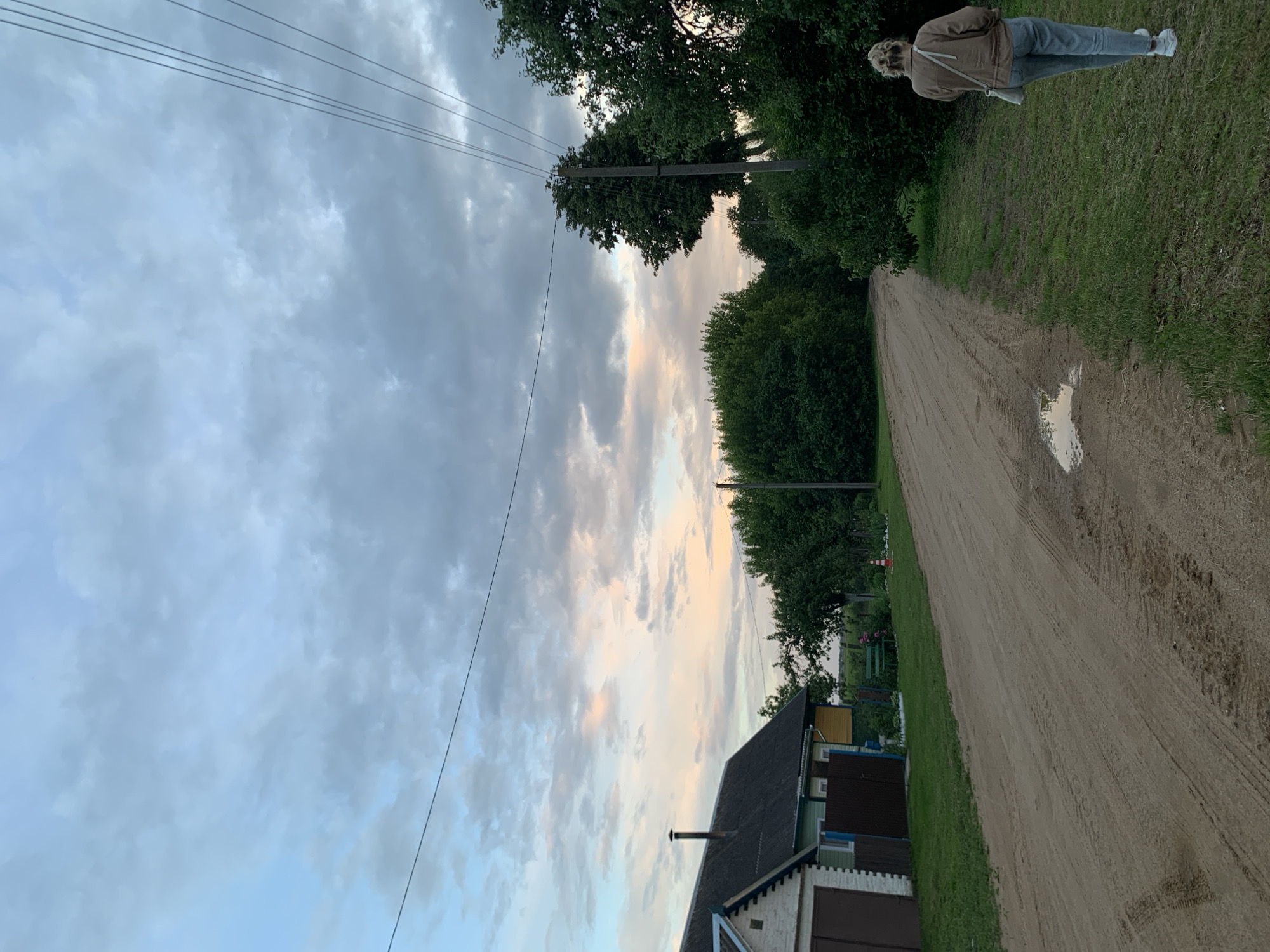 In the village of Bleychitsy, Belarus, at dusk, a woman with blonde hair pulled back in a bun walks away from the camera on the right side of a dirt road. She wears a brown jacket, light blue jeans, and white sneakers, with a light-colored crossbody bag. The road stretches into the distance, showing tire tracks and a small puddle reflecting the sky. On the left side of the road, a house with a white brick facade and a dark roof has a brown garage door. Adjacent to it is a smaller building with green and yellow siding. A few trees are visible behind the house, and a larger cluster of dense green trees forms a wall on the right side of the road, extending into the background. A utility pole with multiple wires stands prominently amidst the trees. The sky above is cloudy, with hues of pale pink and orange from the setting sun peeking through the grey clouds.