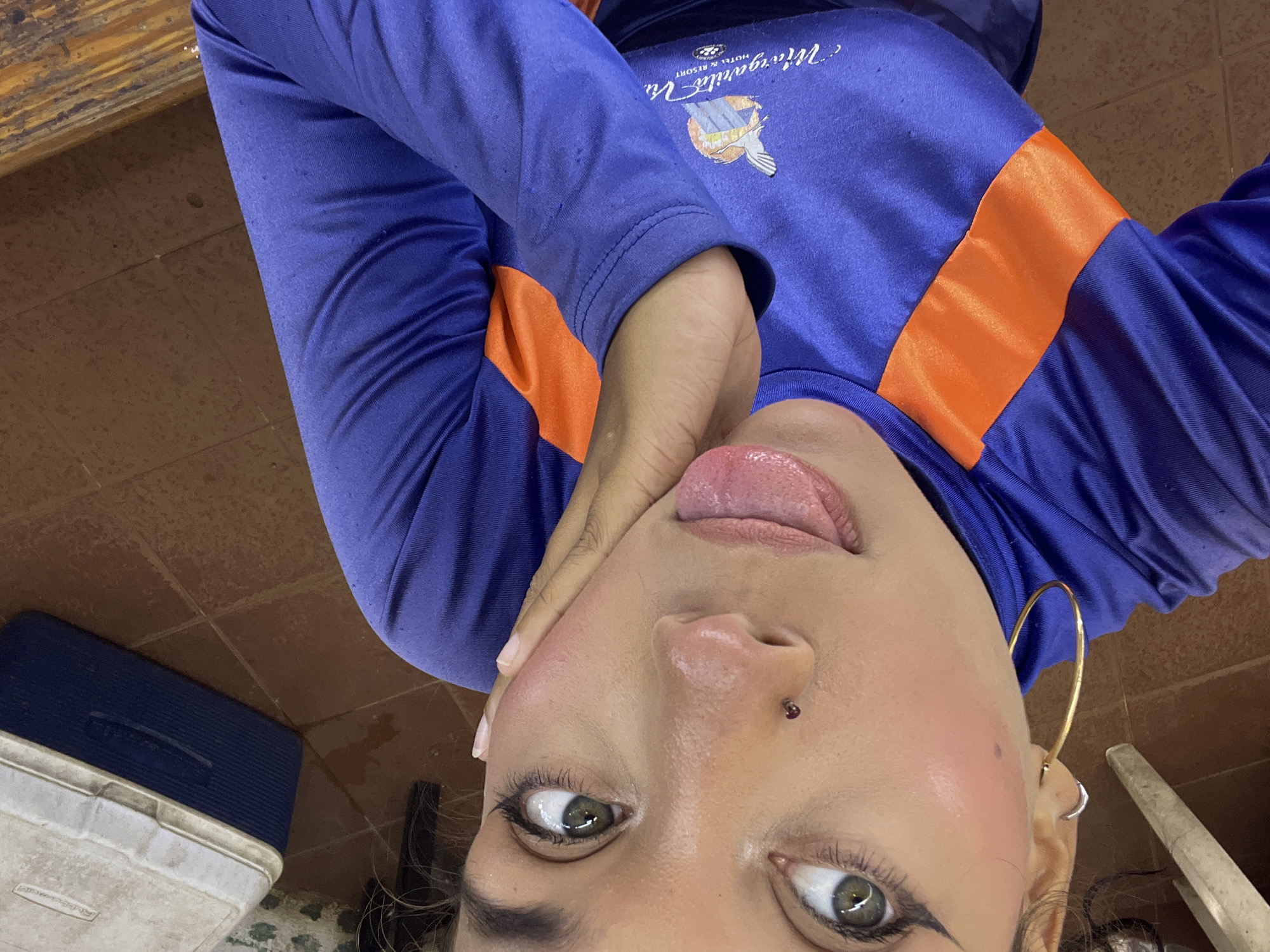 A high-angle, close-up shot captures a young woman in a blue long-sleeved shirt with orange accents, featuring a logo that reads "Margarita Vie Hotel & Resort." She is outdoors, leaning against what appears to be a wooden structure, with a tiled floor and a white cooler visible in the background. The woman has light brown skin, green eyes, and is wearing eyeliner and blush. Her left hand is resting on her cheek, and her tongue is slightly extended. She has a nose ring with a red gem. The overall setting suggests a casual environment in Porlamar, Venezuela.