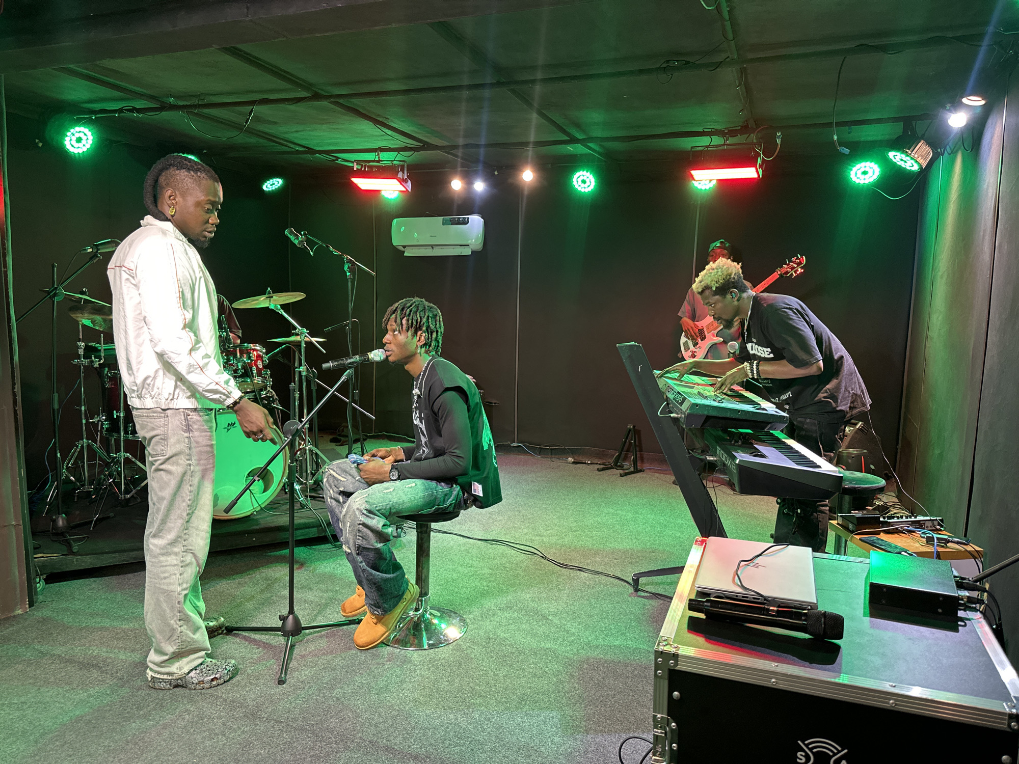 In what appears to be a music studio or rehearsal space in Gwong, Nigeria, four individuals are engaged in a musical activity. The room is dimly lit, with vibrant green and red stage lights illuminating the scene.
On the left, a man stands wearing a white jacket and light-colored jeans. He is facing another man who is seated on a stool in the center of the frame. The seated man is wearing a dark t-shirt with a graphic print, ripped jeans, and yellow shoes. He is looking towards the standing man and has a microphone positioned in front of him. A drum set is visible behind the standing man, suggesting he might be a drummer or involved in that aspect of the music.
To the right of the seated musician, another man is playing a keyboard setup. He has light-colored hair and is wearing a dark t-shirt. Further in the background, a fourth individual is visible holding a red electric guitar.
The environment is equipped for music production and performance, with various microphones, stands, keyboards, a laptop, audio equipment, and a large speaker or flight case on the floor in the foreground. The overall impression is of a band or musical group rehearsing or preparing for a performance.
