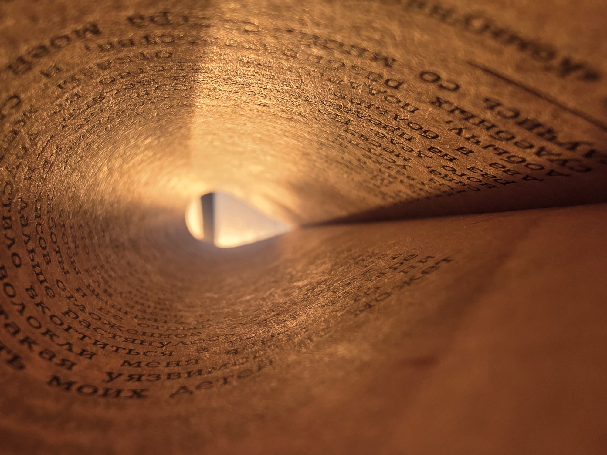 This is a close-up image taken inside a rolled-up book, appearing as if looking through a tunnel. The pages are filled with Russian text. The lighting is warm, with a bright spot in the center where the rolled pages create an opening to the outside. The book appears to have aged, with a slightly yellowed or brownish tone to the paper. The text itself is black and somewhat faded. The location cannot be determined to be in Chaikovsky, Russia, based on the visual information provided.