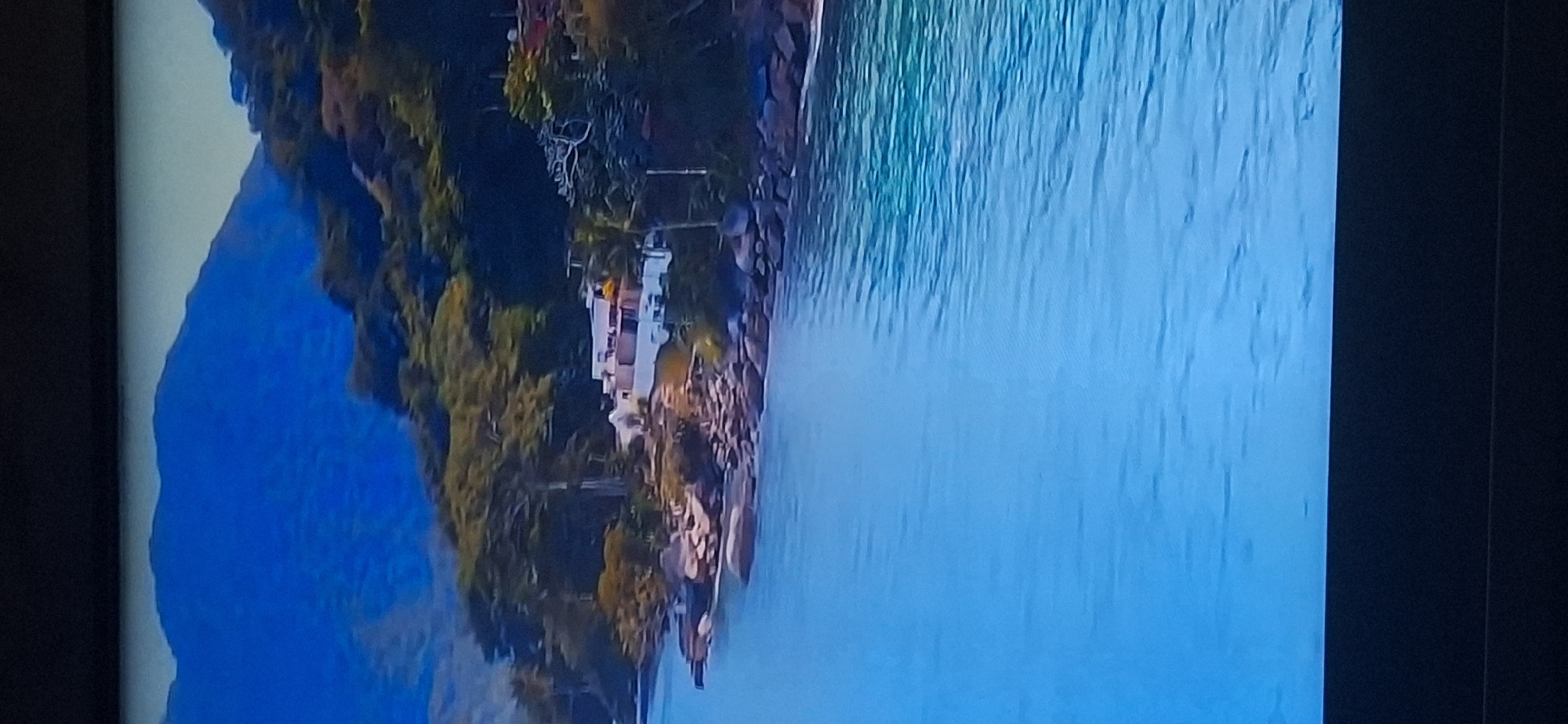 The image depicts a scenic coastal view with a modern white building nestled on a rocky incline overlooking a vibrant blue body of water. Lush green vegetation covers the hillside, with mature trees dotting the landscape. In the background, distant mountains rise, their blue hues fading into the sky. The water is clear and calm, with subtle ripples reflecting the sunlight. There are no people visible in the image. The scene suggests a tranquil and luxurious retreat in a tropical or subtropical environment. The location in Bila Tserkva, Ukraine is not relevant to the visual content of the image.