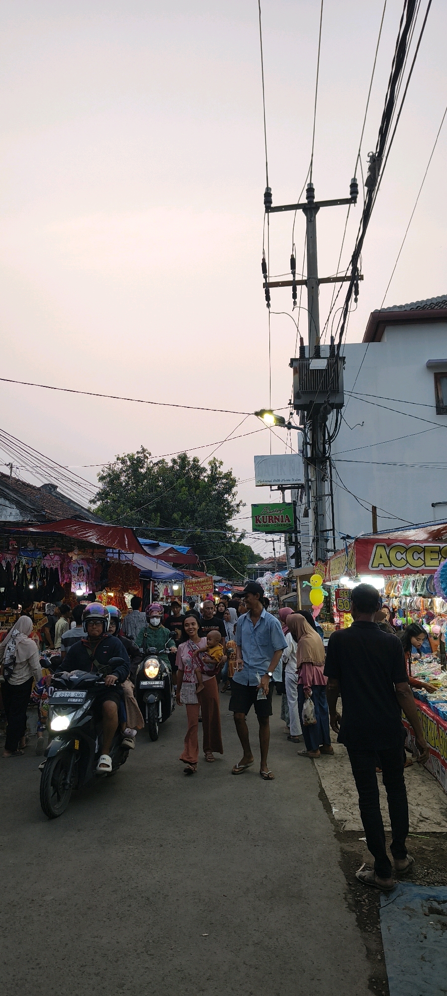 The image depicts a bustling street scene in Trusmi Wetan, Indonesia, during what appears to be an evening market or festival. The sky is a soft, pale orange, suggesting either sunrise or sunset. Power lines and a transformer are prominently visible overhead, with a street lamp illuminating the scene.
The street is lined with market stalls on both sides, covered by red and blue tarps. The stalls are laden with various goods, including clothing, accessories, and potentially food items. People are actively engaged in the market; some are browsing the stalls, while others are walking along the street. A man is riding a motorcycle in the foreground, with another motorcycle behind him carrying passengers. A woman is holding a baby, and a group of people are interacting near the stalls.
The overall atmosphere is one of vibrant activity and community engagement, typical of local markets in Indonesia. The attire of the people, predominantly casual clothing and headscarves for the women, reflects the local culture. The presence of signage, though partially obscured, indicates commercial activity within the area. The ground is paved with asphalt, and the overall setting suggests a well-established, possibly rural or semi-urban, location.