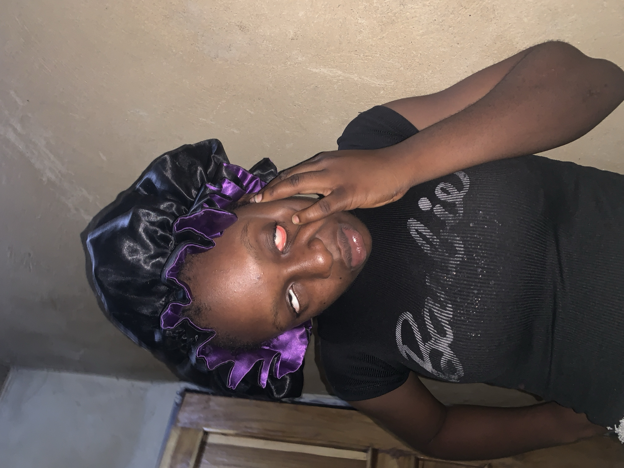A young woman with dark skin and a black bonnet with purple trim is pictured. Her eyes are open and appear red and inflamed. She is wearing a black t-shirt with the word "Barbie" printed in white. Her left hand is touching her face, near her eye. The background is a textured, light brown wall. To the left, a wooden door and frame are visible. This appears to be an indoor scene, possibly a bedroom or private living space. The lighting is dim, casting shadows. The woman's expression is difficult to ascertain due to her eye condition and hand placement. There are no other individuals or objects of note in the immediate vicinity. The context of Agban, Nigeria, is not visually apparent from the image itself.