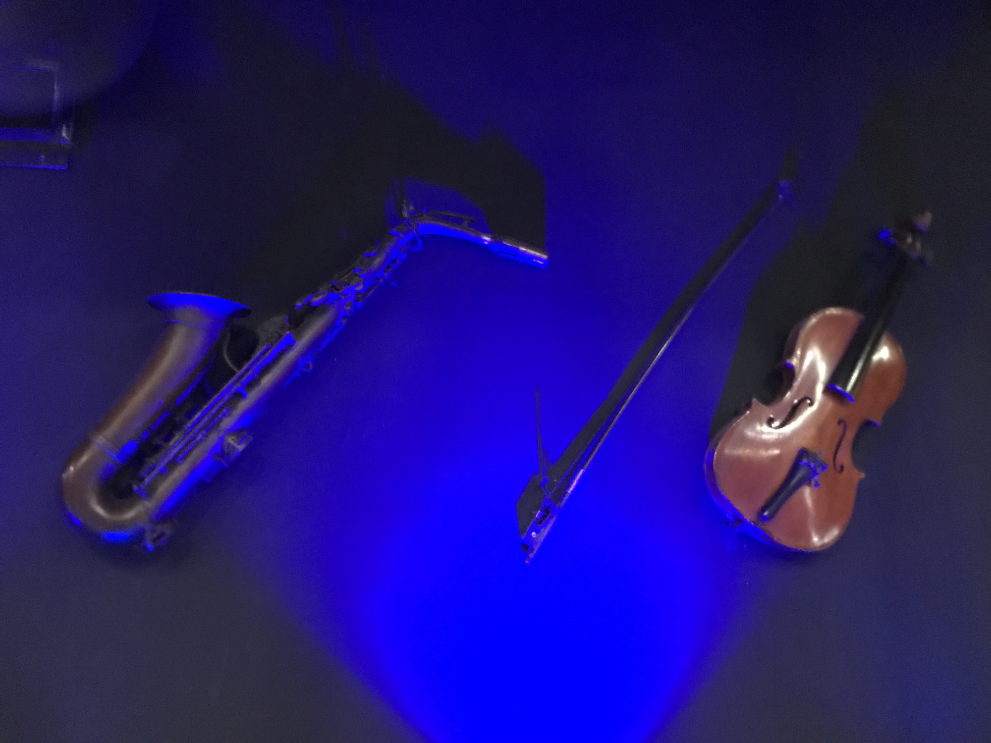 The image shows a saxophone and a violin lying on a dark surface with blue lighting. The saxophone is on the left side of the image and the violin is on the right side with its bow. The background is a dark wall. The location is presumed to be in the city of Medan, Indonesia.