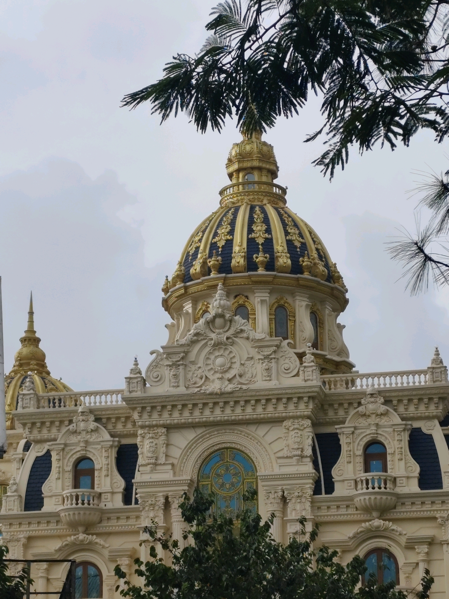 The image captures a detailed view of the exterior of a grand, ornate building, likely a historical landmark or a lavish hotel, under an overcast sky. The architecture is characterized by its elaborate Baroque and Rococo influences, featuring intricate carvings, decorative moldings, and a prominent golden dome with dark blue accents. The building's facade is cream-colored and adorned with balconies, arched windows, and balustrades. In the foreground and framing the top of the image, lush green foliage from trees partially obscures the view, suggesting an outdoor setting.
This particular building is known as the "Crazy House" or "Hang Nga Guesthouse" in Da Lat, Vietnam. It is an architectural marvel designed by Vietnamese architect Đặng Việt Nga, known for its whimsical and organic design that deviates from conventional architecture. The building's unconventional shape and construction are inspired by nature, with winding staircases, caves, and surreal elements integrated throughout.