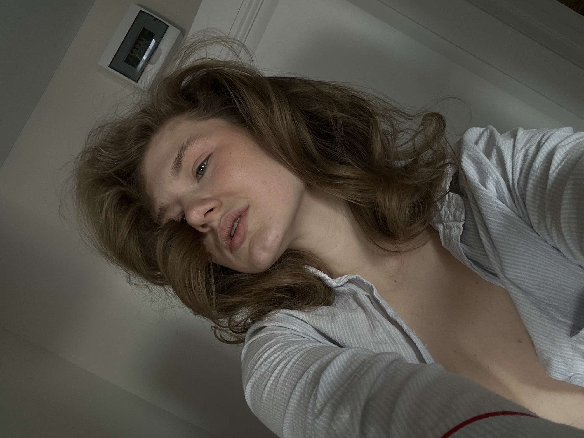 A low-angle, medium close-up shows a fair-skinned person with shoulder-length wavy brown hair and blue eyes. The person is wearing a striped, collared shirt, unbuttoned to reveal the upper chest. The background is a plain off-white wall. A digital clock is mounted on the wall. There is no indication of the location being Kazan, Russia.