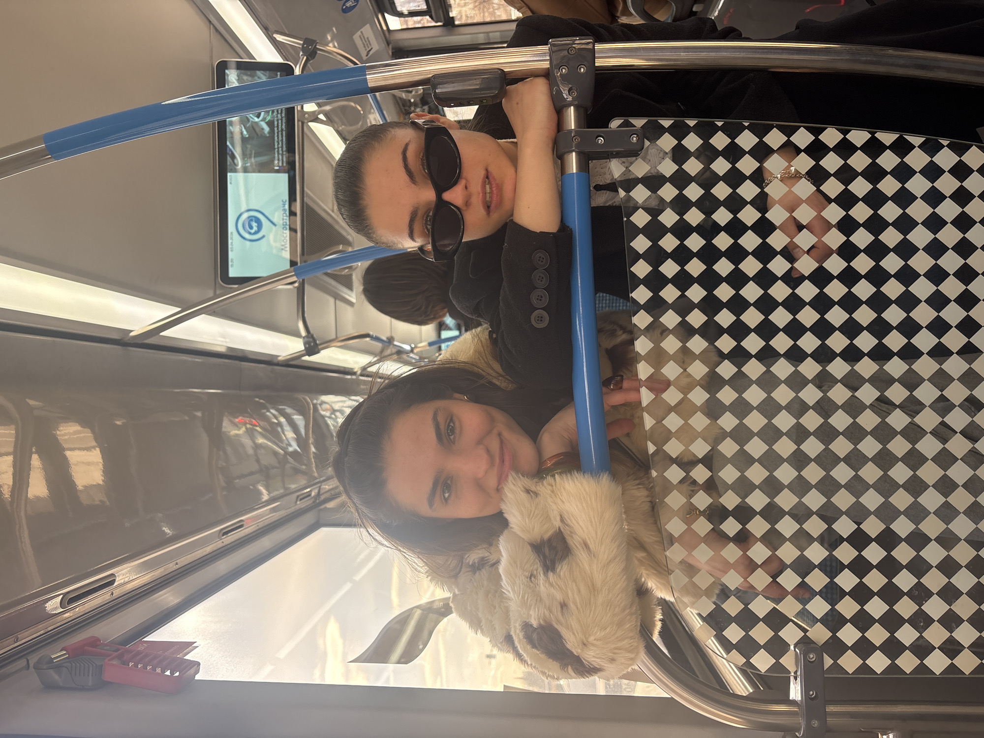 Two young women are on a Moscow Metro train. One woman wears sunglasses and a black jacket with buttons. The other woman wears a coat with fur trim. They are holding onto the blue handrails. There is a digital information display with Russian text, and a checkered pattern on the train's interior. A hand holding a silver bracelet rests on the checkered surface. Tools are on a ledge.