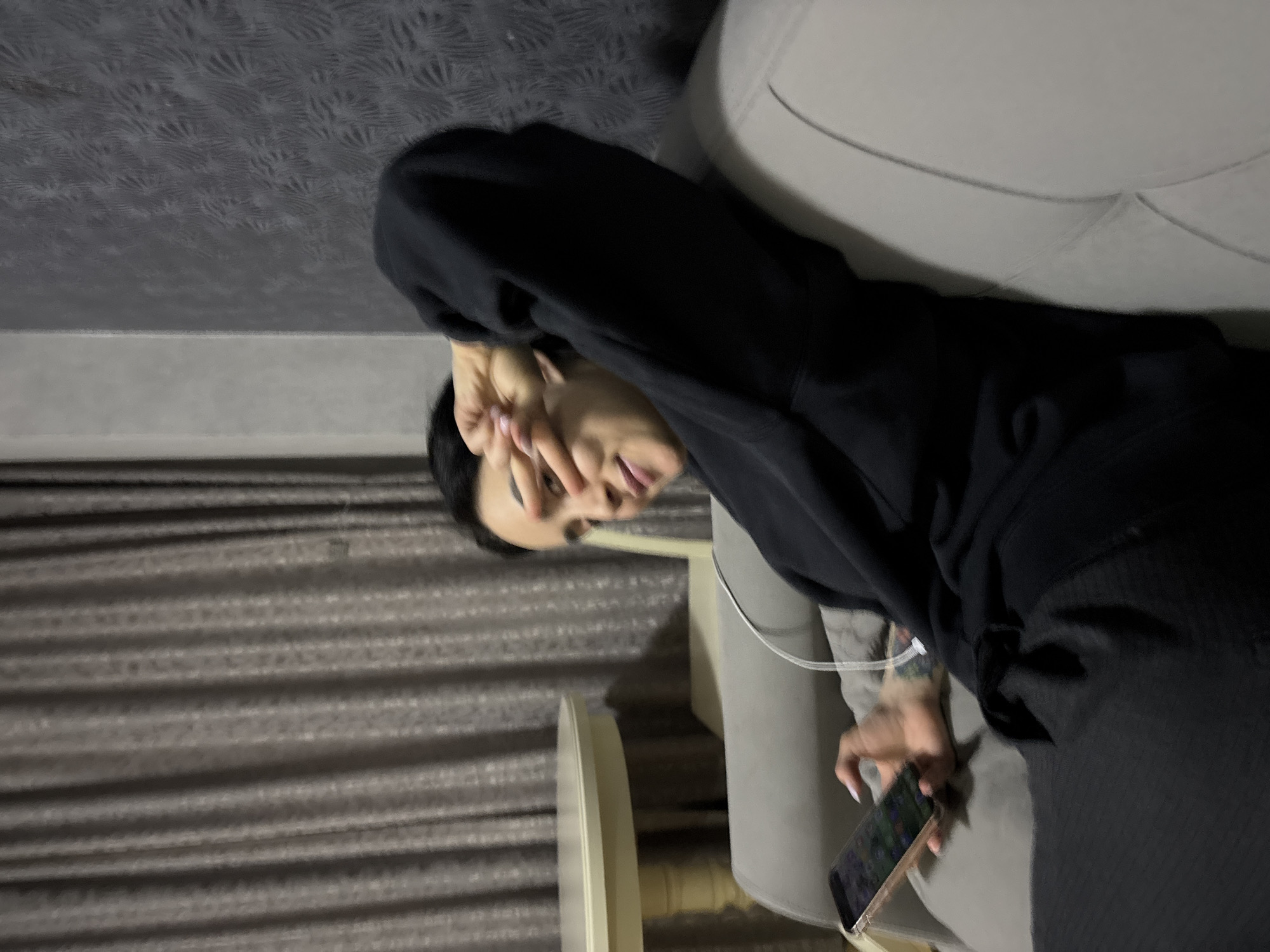 An indoor scene, when correctly oriented, depicts a person lounging on a light grey upholstered couch, likely in a living room or lounge. The individual has dark hair and is dressed in a black hooded sweatshirt. Their right hand is raised, partially covering their face and eyes, with a hint of their tongue visible. Their left hand holds a light-colored smartphone, possibly a gold or rose gold model, to which a white cable (either for charging or headphones) is connected. The person has light pink nail polish on their fingernails, and a blurry tattoo is visible on their left forearm.
The background features a dark grey wall adorned with a patterned wallpaper, reminiscent of fern leaves or feathers. The floor is covered with a dark, ribbed material, possibly carpet. A small, light-colored round or oval table is partially visible in the foreground. The overall lighting is dim, suggesting it is evening or nighttime. No specific location cues are discernible within the image itself, though the context provided indicates Tbilisi, Georgia. No visible text can be clearly read on the phone screen or elsewhere in the image.