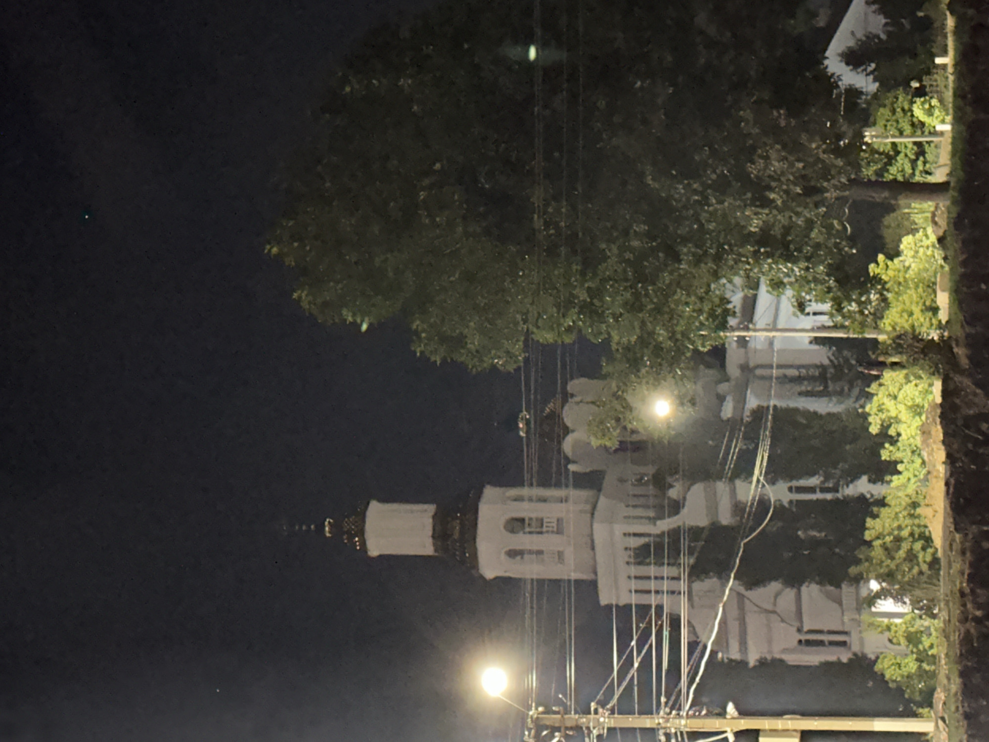 An image captured at night shows a white church with a tall bell tower, its top section adorned with decorative gold accents. The church is illuminated, highlighting its architectural details. In the foreground, a large, leafy tree partially obstructs the view of the church. Utility poles and power lines are visible to the left of the church, and a bright street lamp illuminates the area. The sky above is dark, with a single faint green light visible in the upper right quadrant. The ground in the foreground appears to be a construction or excavation site with exposed earth and some low-lying vegetation. The overall scene suggests an urban environment in Nevinnomyssk, Russia, likely a historical or religious site on display at night.