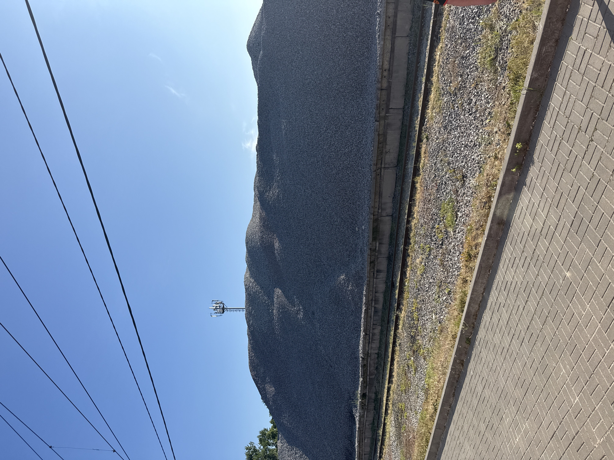 The image depicts a large pile of crushed stone situated next to a railway track. A cell tower is visible on top of the pile. The scene is outdoors, under a clear blue sky. In the foreground, a paved walkway is present, with a concrete curb separating it from the railway line and the crushed stone. Power lines stretch across the upper portion of the image. No people are clearly visible in the frame. The location is identified as Arzamas, Russia.
