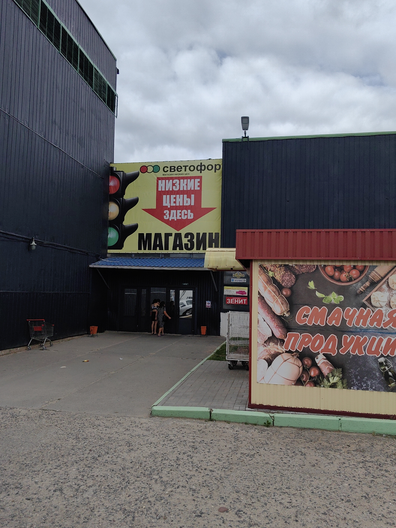 The image captures the exterior of a building in Pastavy, Belarus. The building features a dark, corrugated metal facade. To the left, a large, bright yellow banner advertises a store named "СВЕТОФОР" (Svetofor), which translates to "Traffic Light." The banner prominently displays a graphic of a traffic light with red, yellow, and green signals, and text reading "НИЗКИЕ ЦЕНЫ ЗДЕСЬ" (Low Prices Here) and "МАГАЗИН" (Store).
To the right of the main building, a smaller, red-roofed structure houses another advertisement featuring images of various meat products, sausages, tomatoes, and herbs, with the text "СМАЧНАЯ ПРОДУКЦИЯ" (Delicious Products) in large, bold, red lettering.
In front of the main building's entrance, two women are visible standing near the doorway. A shopping cart is positioned to the left of the entrance. The ground is paved with asphalt and gravel. The sky is overcast with clouds.