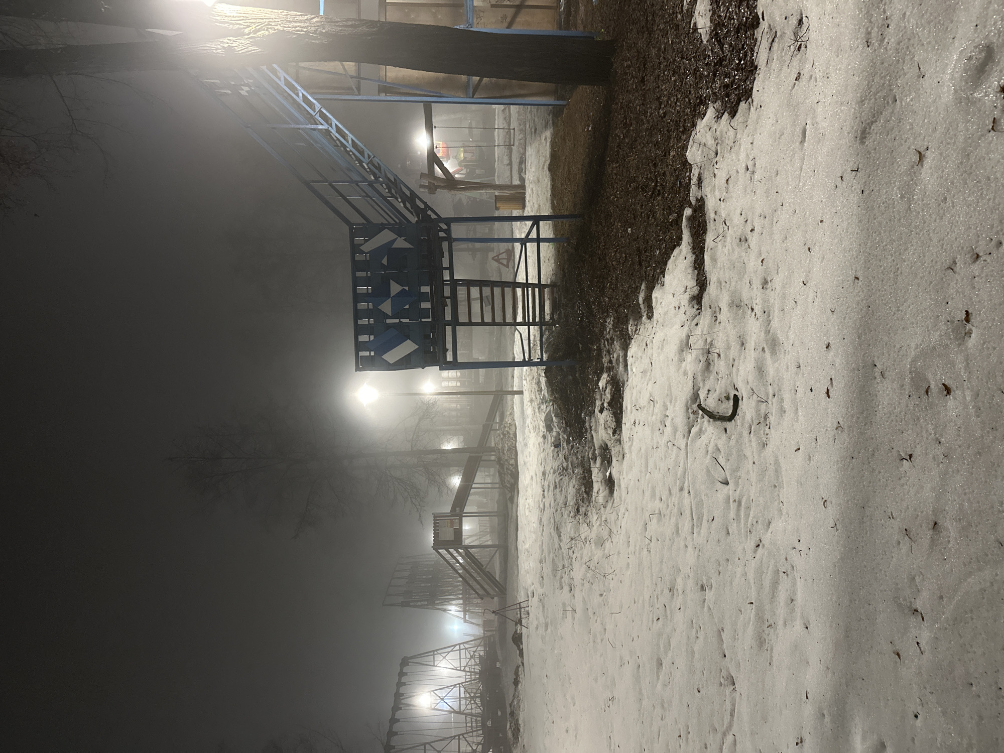 The image depicts a foggy night scene in what appears to be a playground in Sarapul, Russia. Metal playground structures are visible, illuminated by streetlights that are diffused by the heavy fog. Patches of snow cover the ground on one side of the frame, next to a strip of dark ground. No people are visible.