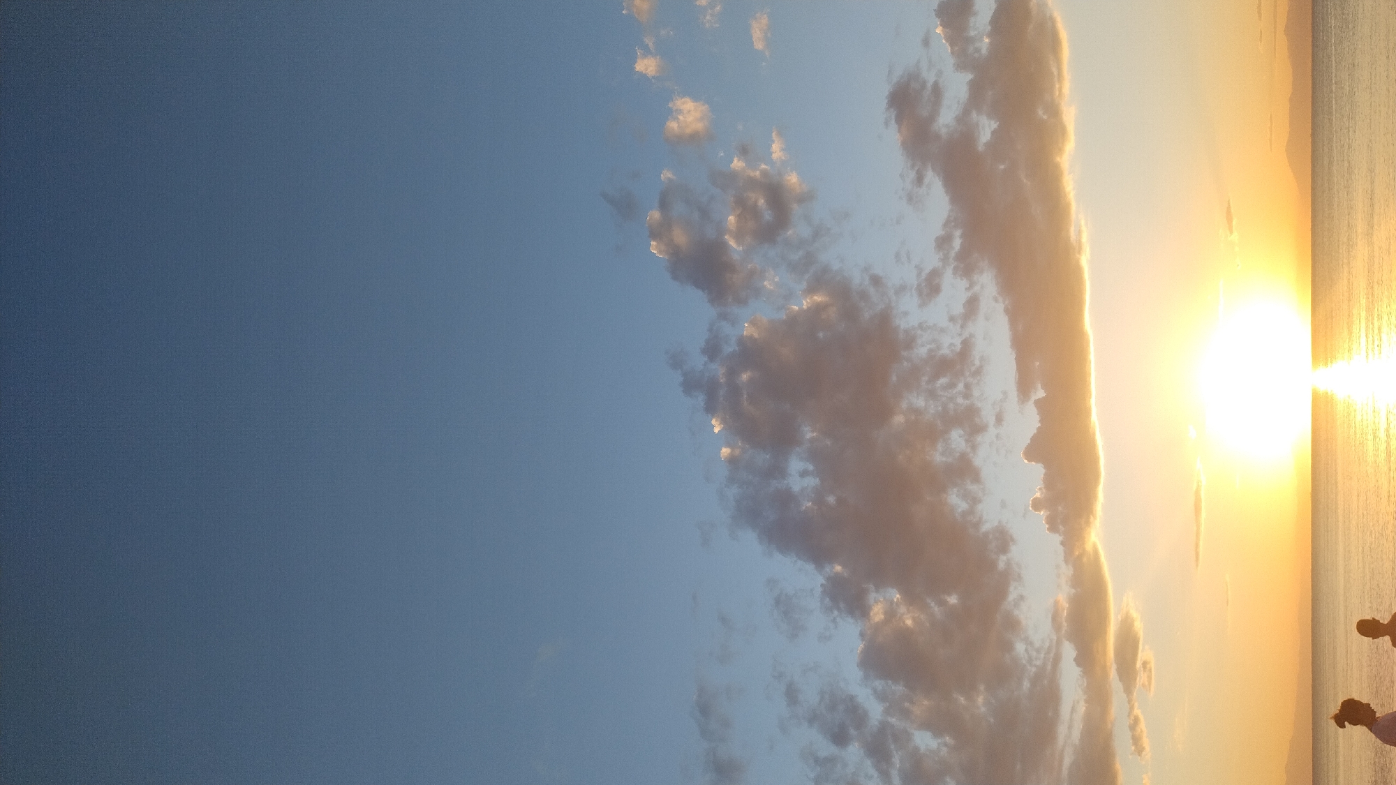 This is a vertical, eye-level, outdoor photograph taken at sunset in Perea, Greece. The scene depicts the sky transitioning from a clear blue at the top to hues of orange and yellow near the horizon, illuminated by the setting sun. Scattered clouds, tinged with the warm colors of sunset, are present in the middle and upper portions of the frame. The lower part of the image shows a calm body of water, likely the sea, reflecting the vibrant colors of the sky. In the foreground, near the left edge of the frame and partially obscured, are two individuals standing by the water. The person on the left is a female with dark hair styled in a bun. The person on the right appears to be a child or a smaller individual. They are facing away from the camera, looking towards the water and the sunset. The overall atmosphere is serene and peaceful, characteristic of a sunset over the sea.